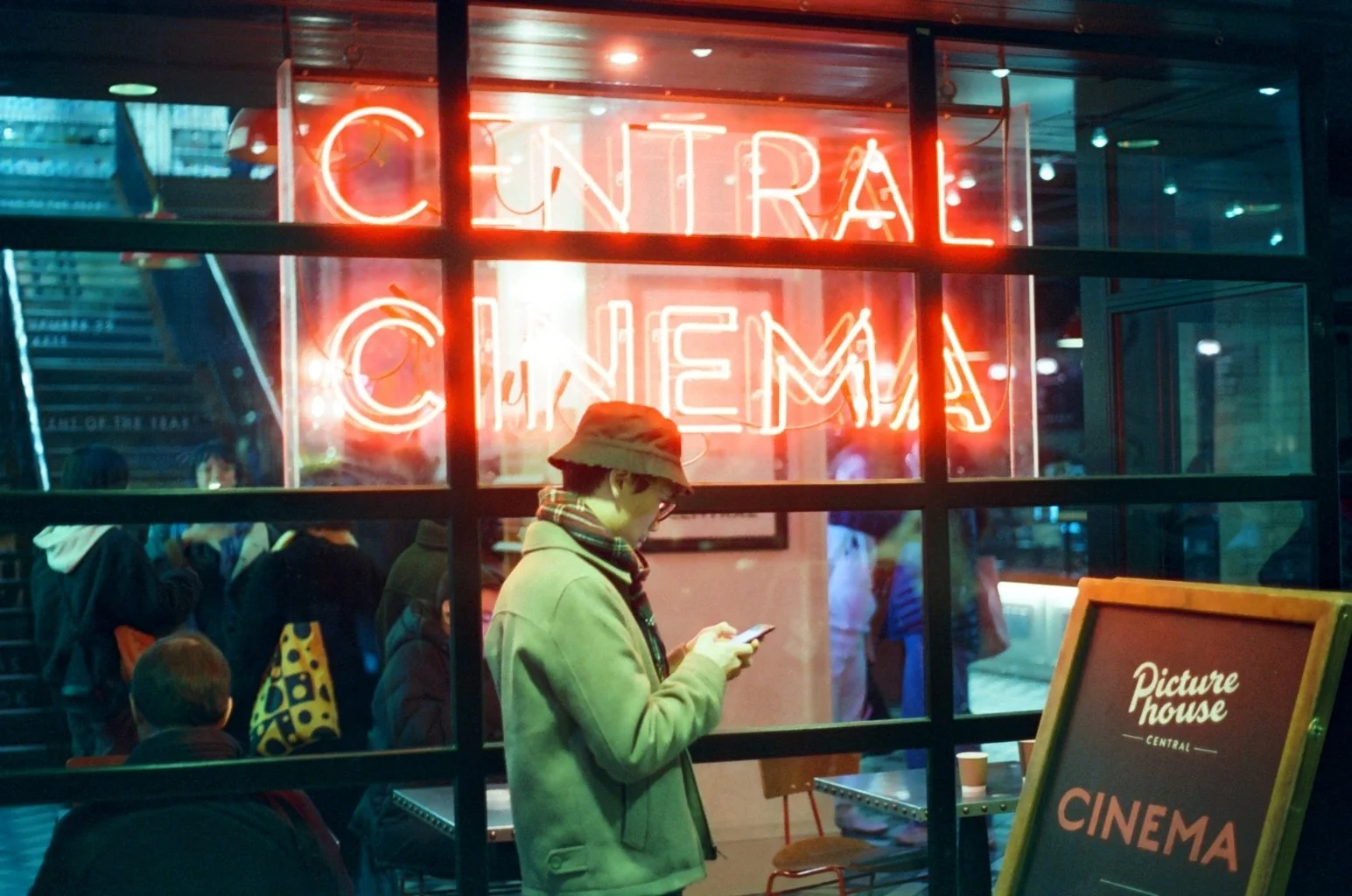 View through window showing a person standing inside a cinema, looking at their phone, with a neon sign that says 'Central Cinema' in the background, and a signboard outside reading 'Picture House Central Cinema'.