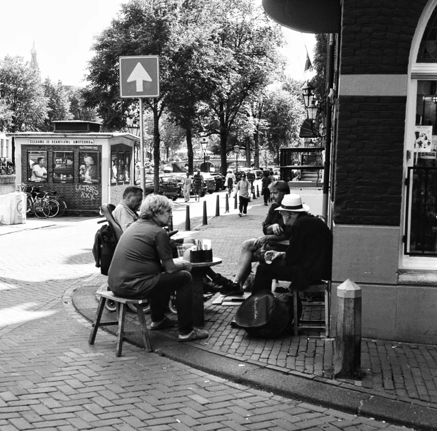 People sitting at an outdoor cafe on a city sidewalk, with trees, cars, and a traffic sign in the background in black and white.
