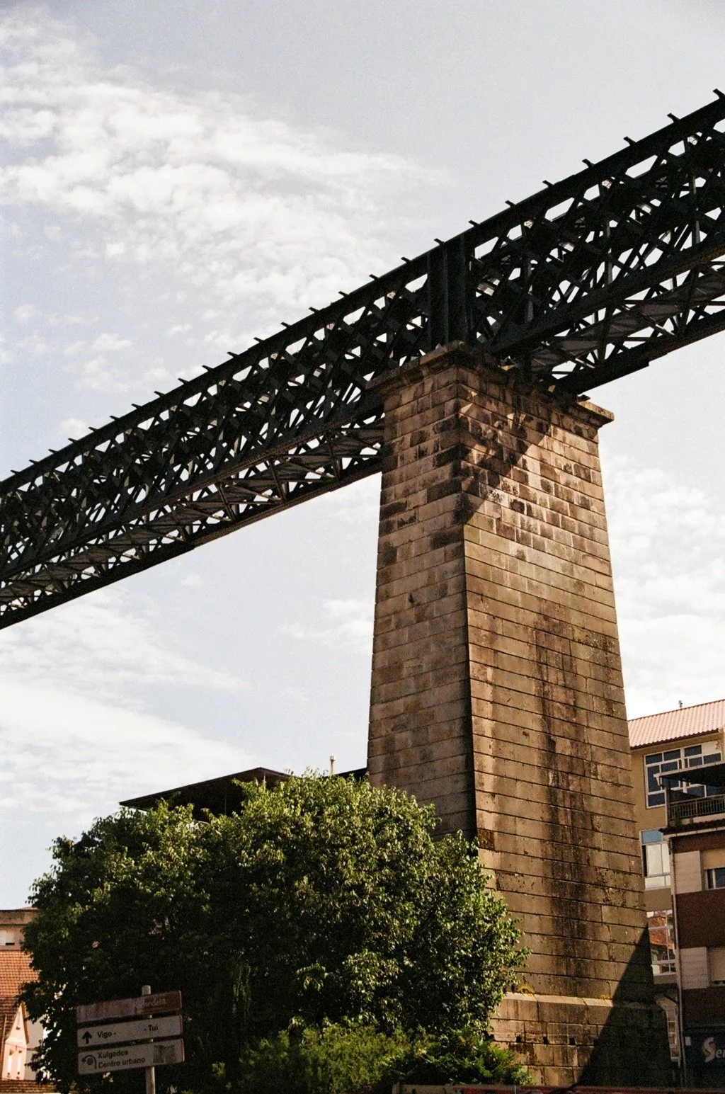 A brick pillar supporting a metal bridge structure against a partly cloudy sky, with trees and buildings in the background.