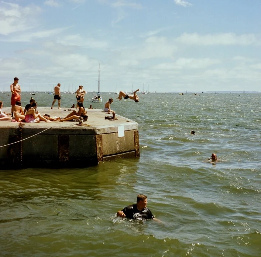 People at a beach enjoy swimming and jumping into the water from a concrete pier on a partly cloudy day, with sailboats in the distance.