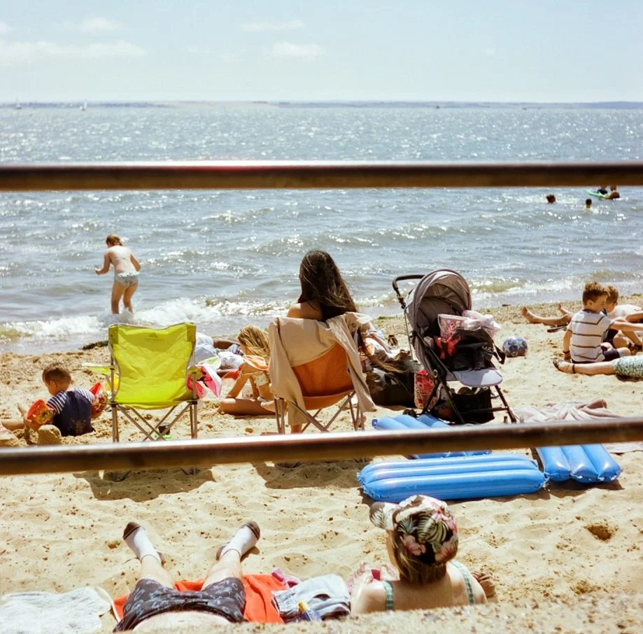 People relaxing and playing on a sandy beach near the ocean, with some sitting on chairs, some lying on towels, and children playing in the water.