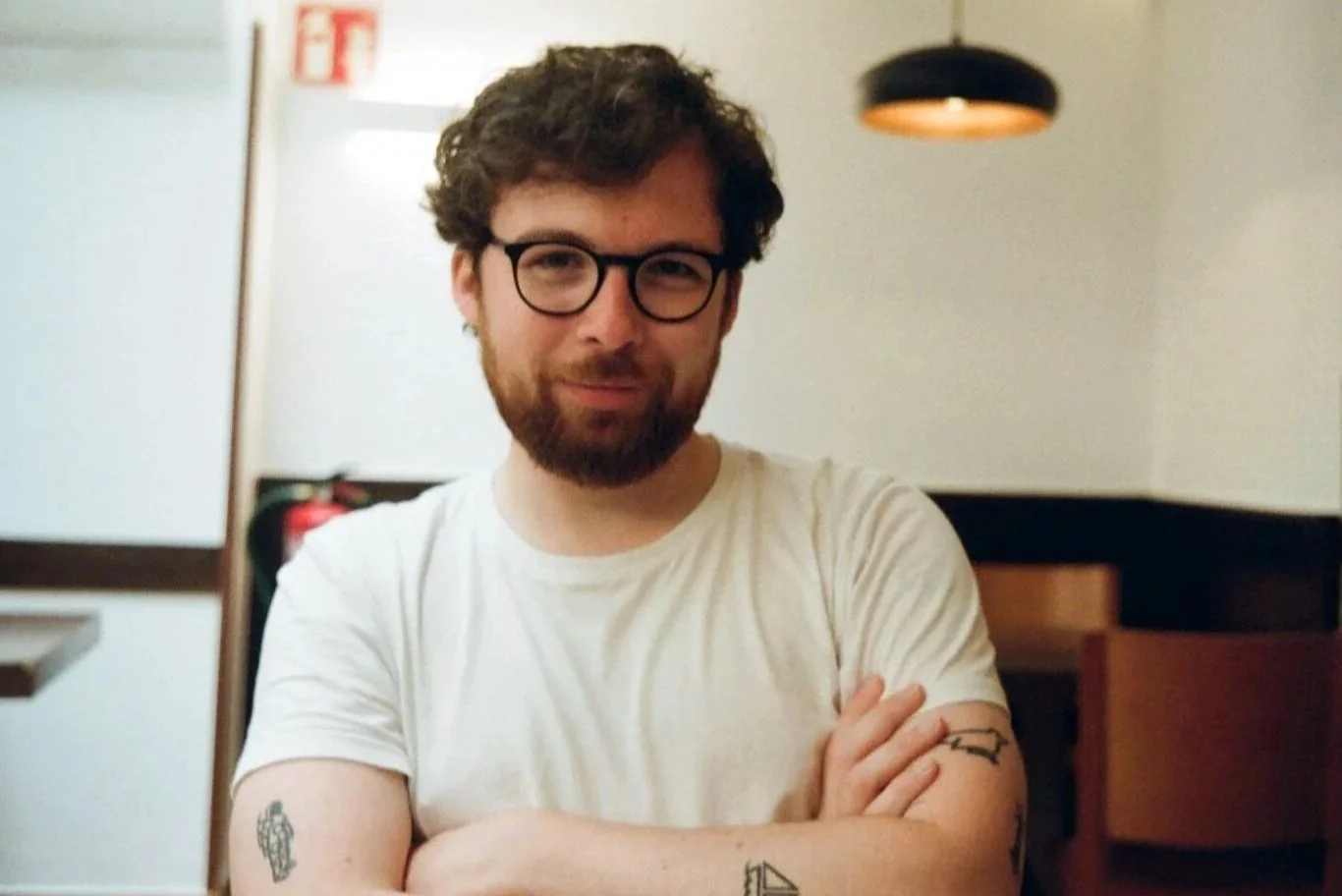 A young man with curly hair, glasses, and a beard sitting with his arms crossed, smiling slightly, wearing a white t-shirt, in an indoor setting with soft lighting.