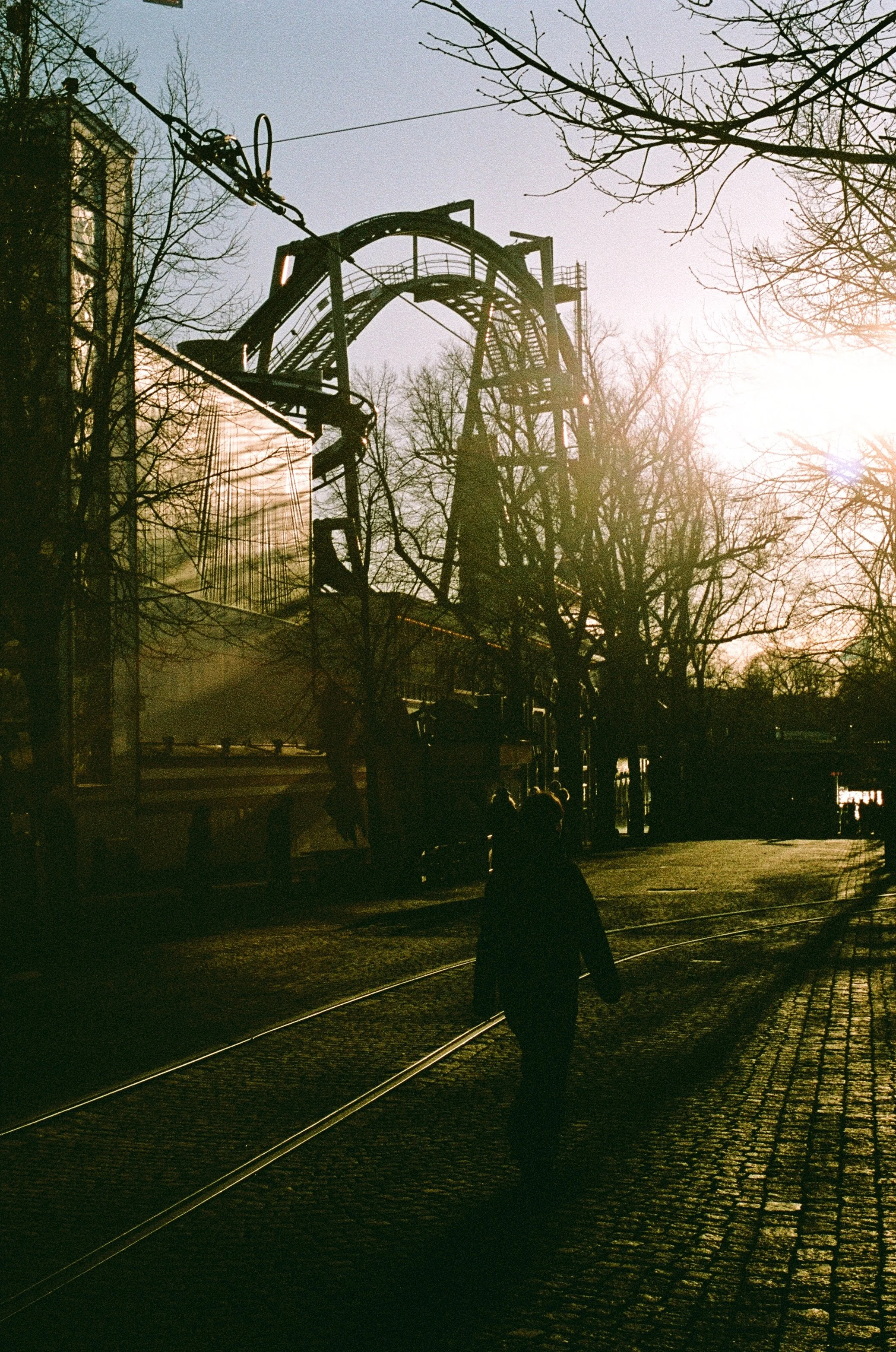 Person walking along tram tracks with amusement park ride in the background during sunset, leafless trees visible.