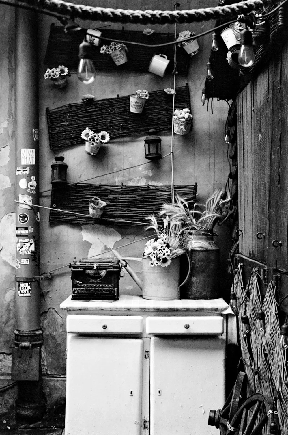 Black and white photo of a vintage kitchen corner with a white cabinet, a potted plant with flowers, wall decor including wooden slats, potted flowers, hanging light bulbs, and a stack of straw-like mats.