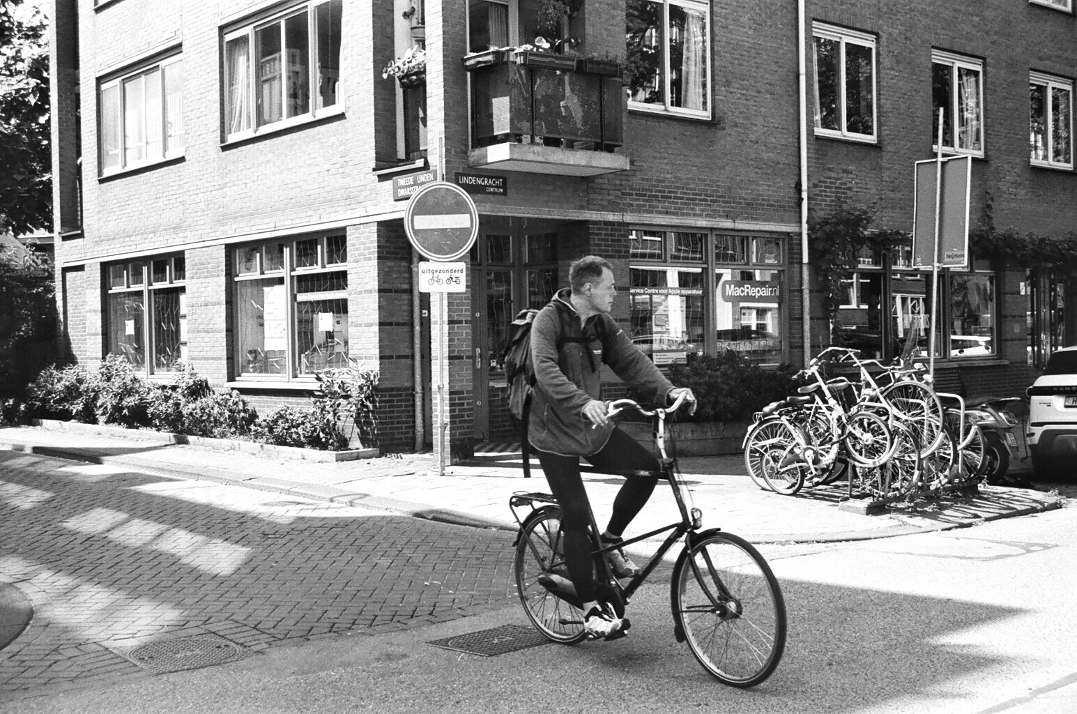 A man riding a bicycle on a city street, with bicycles parked on the sidewalk, a building with large windows, and a street sign visible in the background.