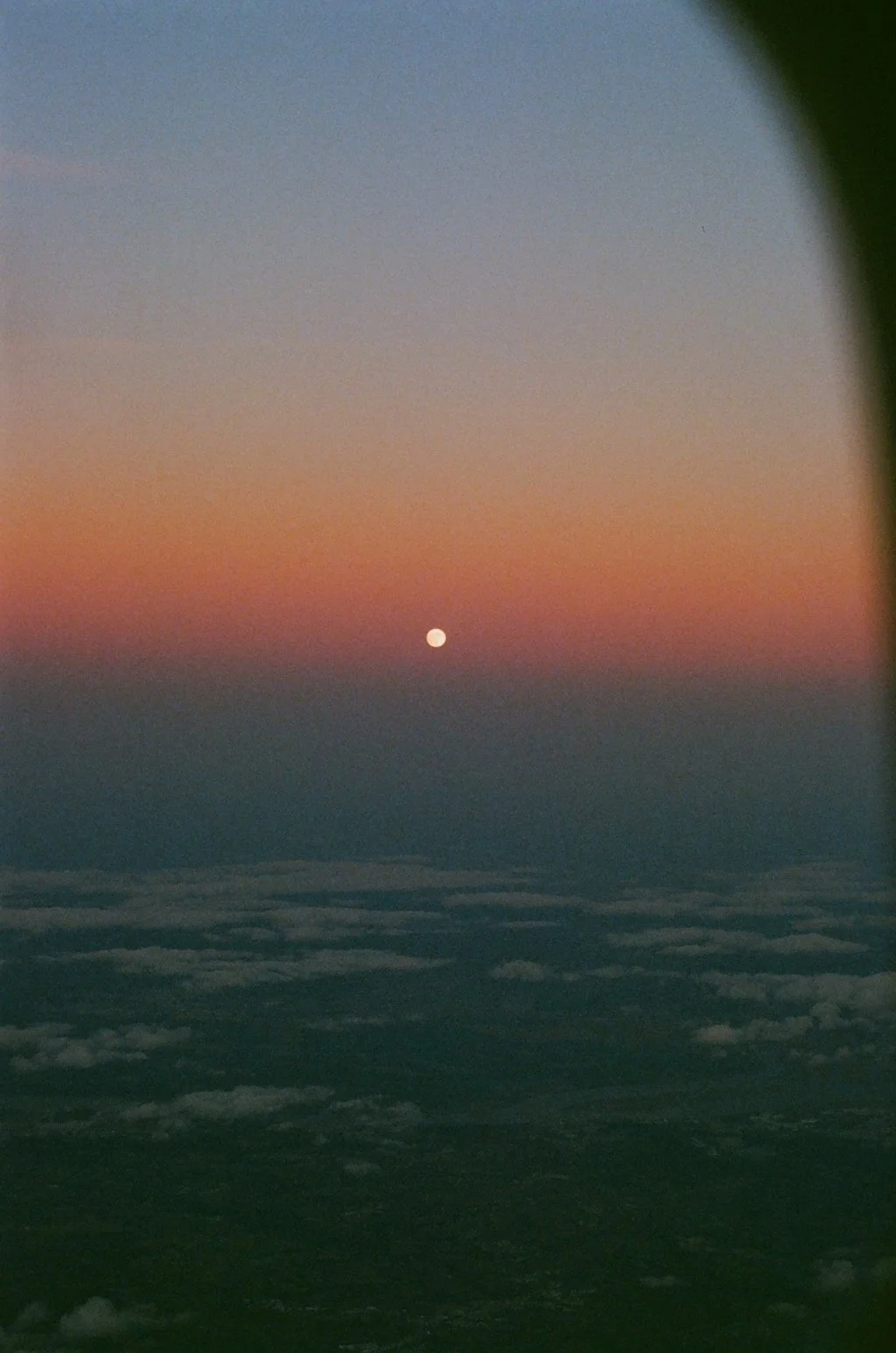 Night sky viewed from an airplane window with clouds below and a small bright moon or planet on the horizon, partially obscured by the airplane window frame.