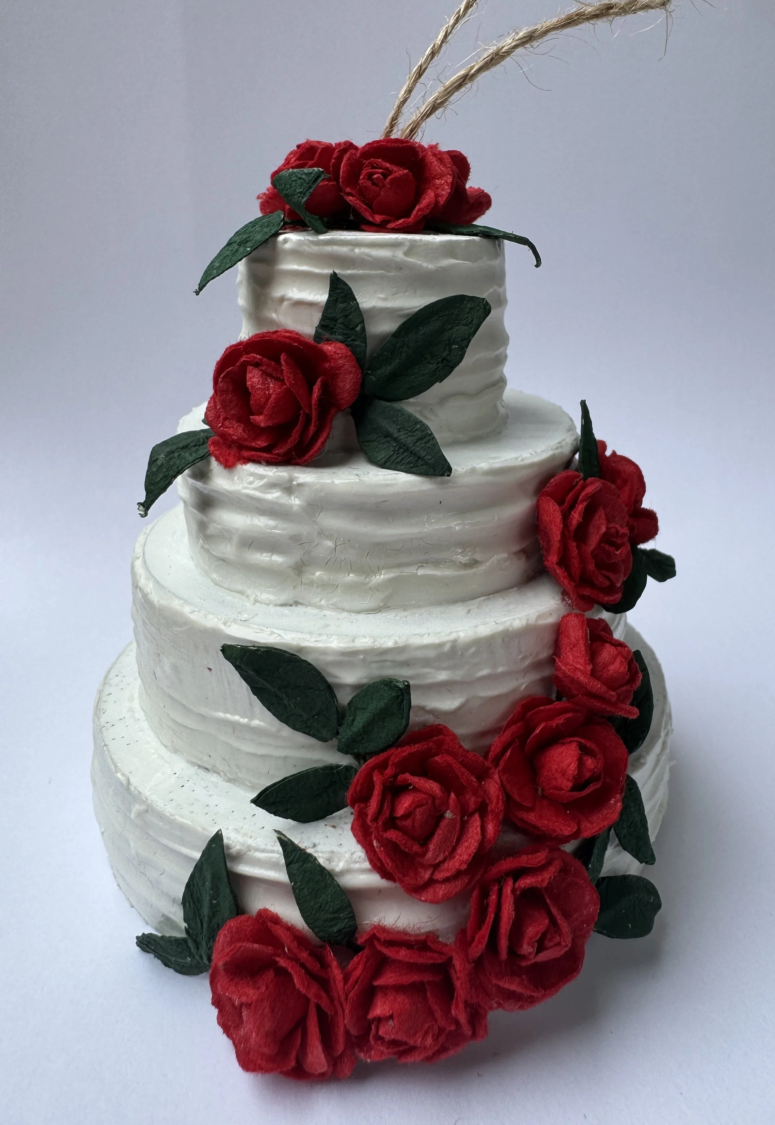 A three-tiered white wedding cake decorated with red roses and green leaves, with a natural twine topper against a plain background.