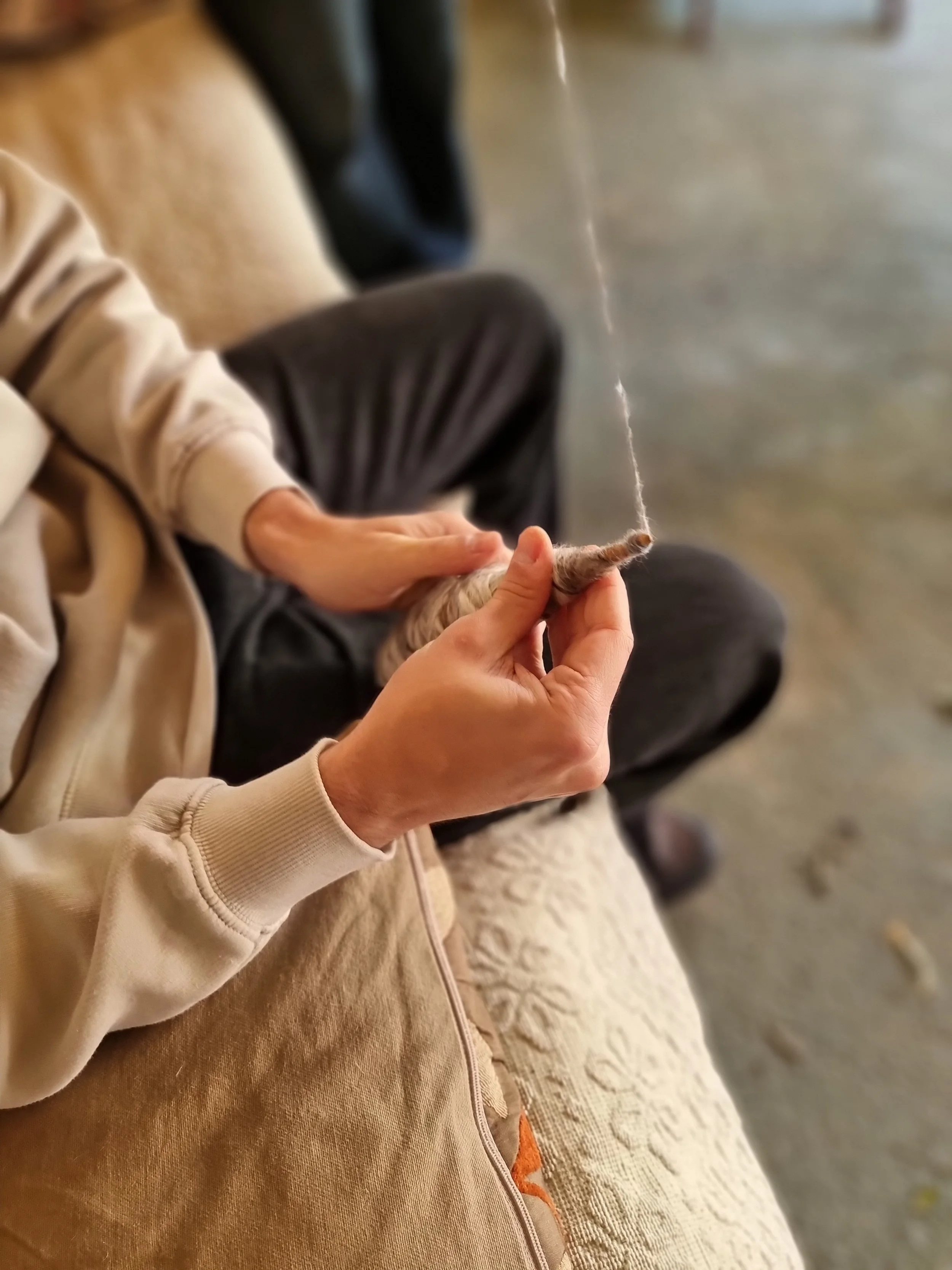 Person knitting with yarn, sitting on a beige textured blanket, and wearing a beige hoodie and brown pants.