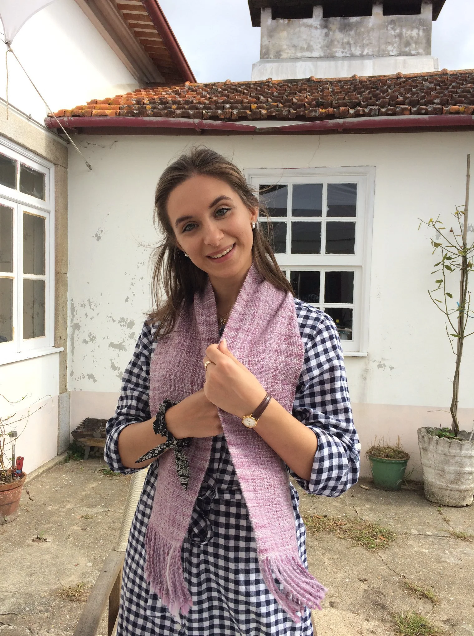 A woman with brown hair, wearing a checkered dress and a hand knitted pink scarf, standing outside in front of a house with white walls, windows, and a tiled roof. She is smiling and holding her scarf with both hands. Made at Monte Frio Alpacas. 