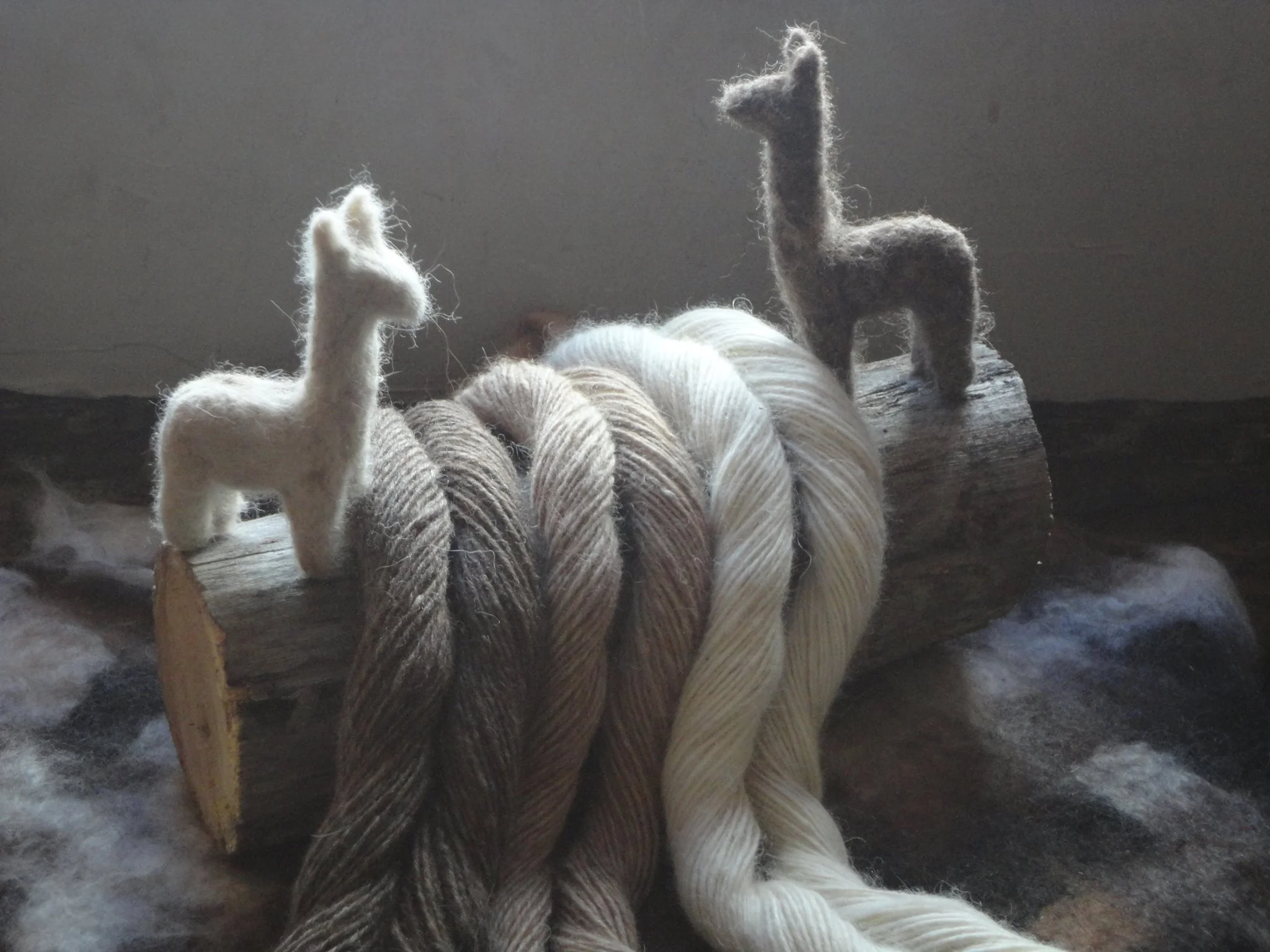 Two hand felt alpaca sculptures standing on a wooden base with yarn coils in front of them, set against a gray background. Made at Monte Frio Alpacas.