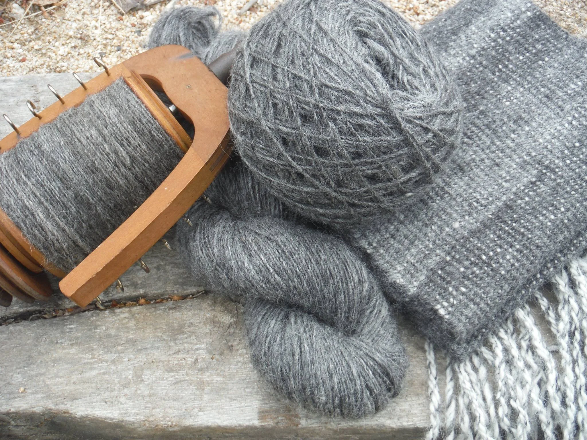Close-up of grey yarn wound into a ball, lying on a grey woven fabric. A wooden knitting loom is partially visible in the scene. Made at Monte Frio Alpacas.