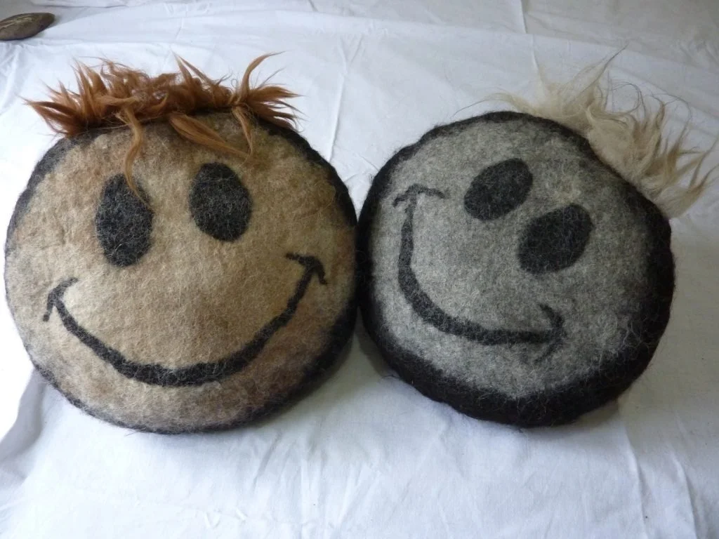 Two round felt pillows with smiley face designs and fuzzy hair, one with brown hair and the other with blond hair, resting on a white surface. Made at Monte Frio Alpacas.