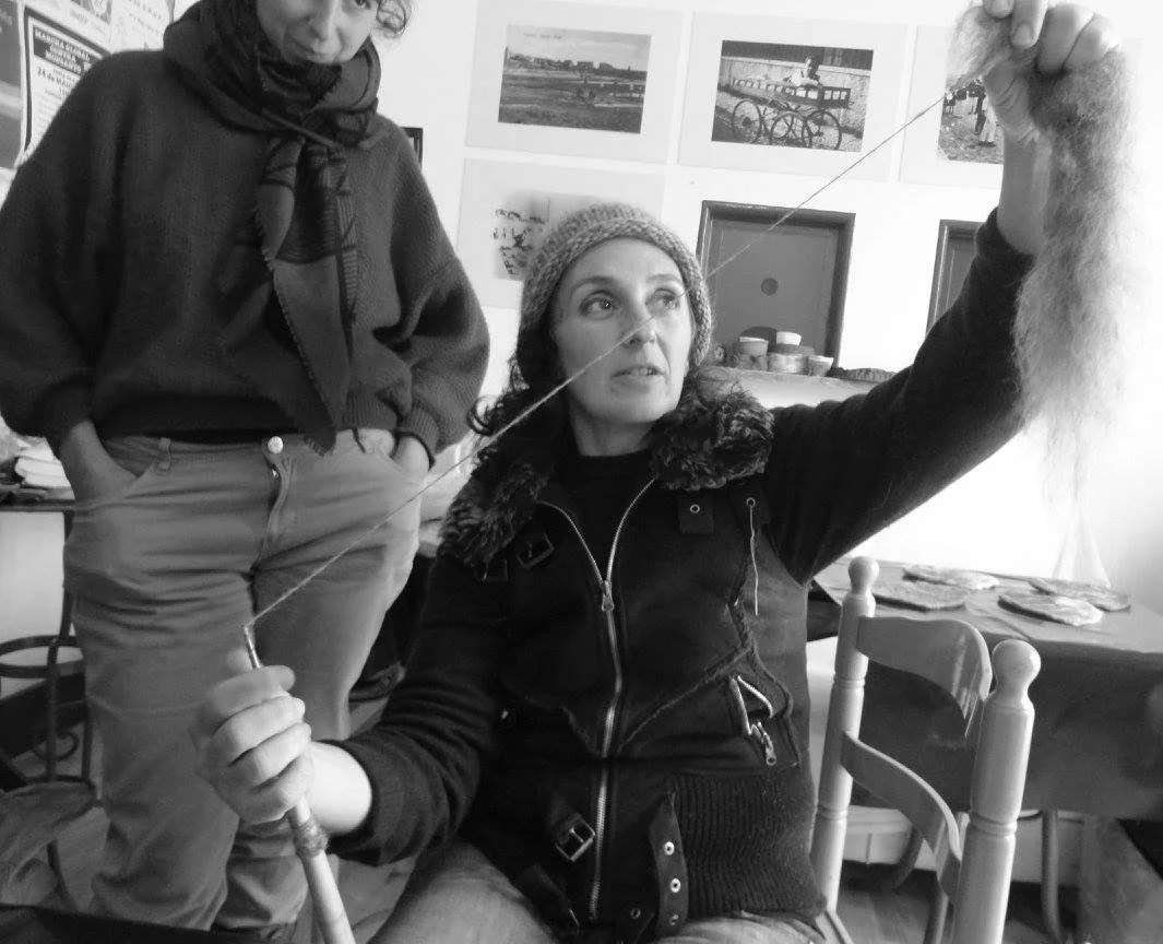 A woman sitting at a table holding a spindle and wool during an activity at Monte Frio Alpacas. She is wearing a knitted hat and a dark jacket, and is focused on the spinning process. The background shows framed photographs.