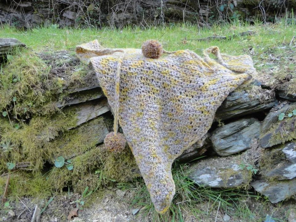 A multicolored crocheted blanket with pom-poms draped over a moss and grass-covered stone wall outdoors. Made at Monte Frio Alpacas.
