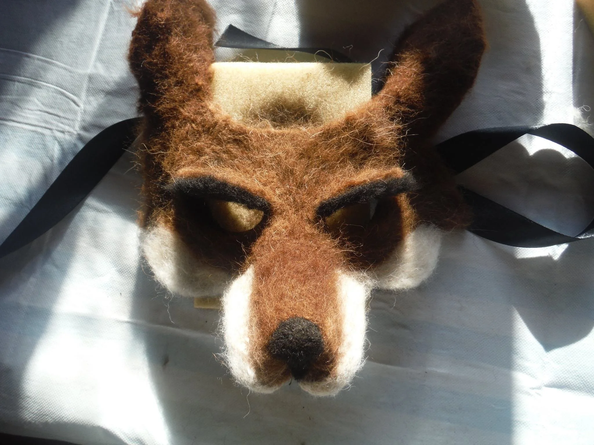 Close-up of a hand felt 3D animal head, possibly a fox or dog, with brown, black, and white details, lying on a light-colored fabric surface. Made at Monte Frio Alpacas. 