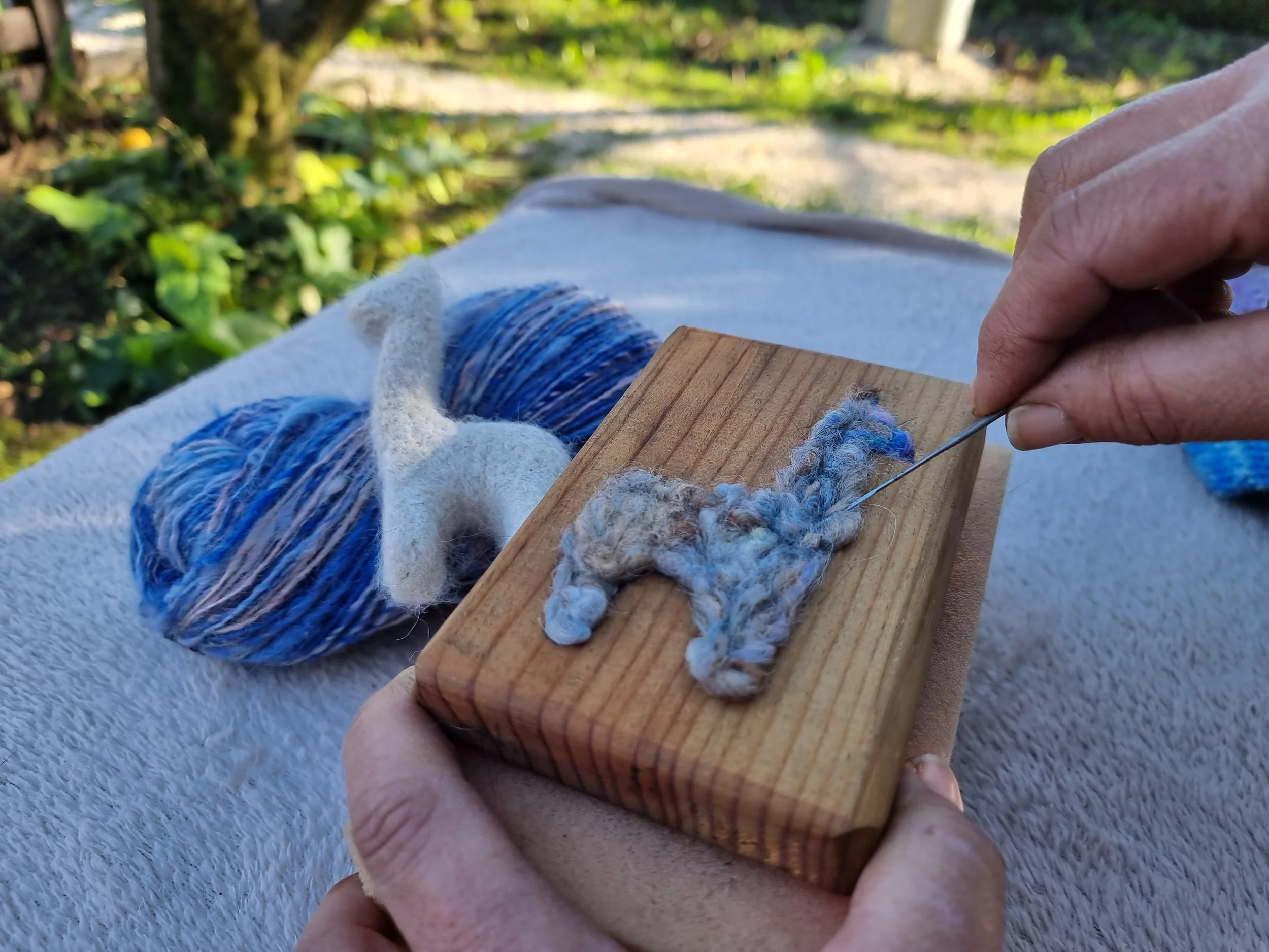 A person's hand holding a wooden block with a piece of yarn wrapped around it for a felting workshop, outdoors with greenery in the background. Made at Monte Frio Alpacas. 