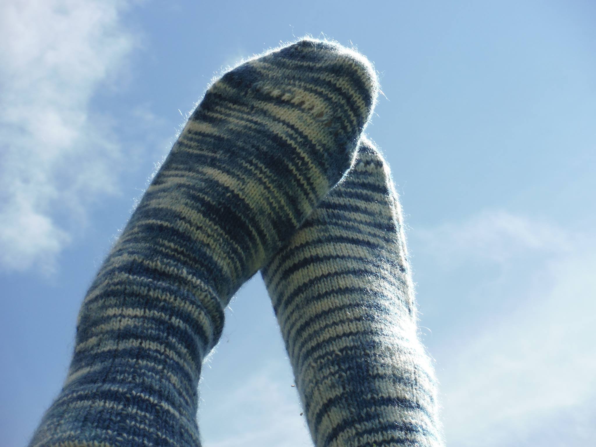 Close-up of a person's leg and foot covered in a knitted sock against a bright blue sky with some clouds. Made at Monte Frio Alpacas. 