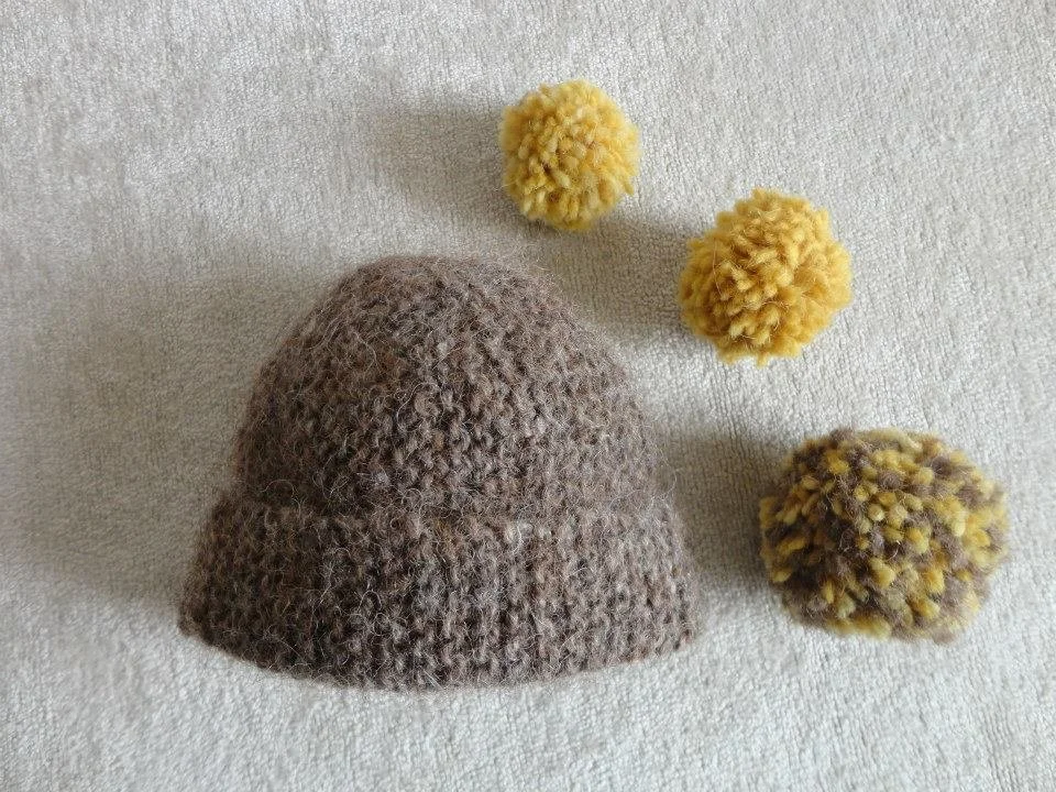 A brown fuzzy knit hat with two yellow pom-poms, and two multicolored pom-poms on a light fabric background. Made at Monte Frio Alpacas. 