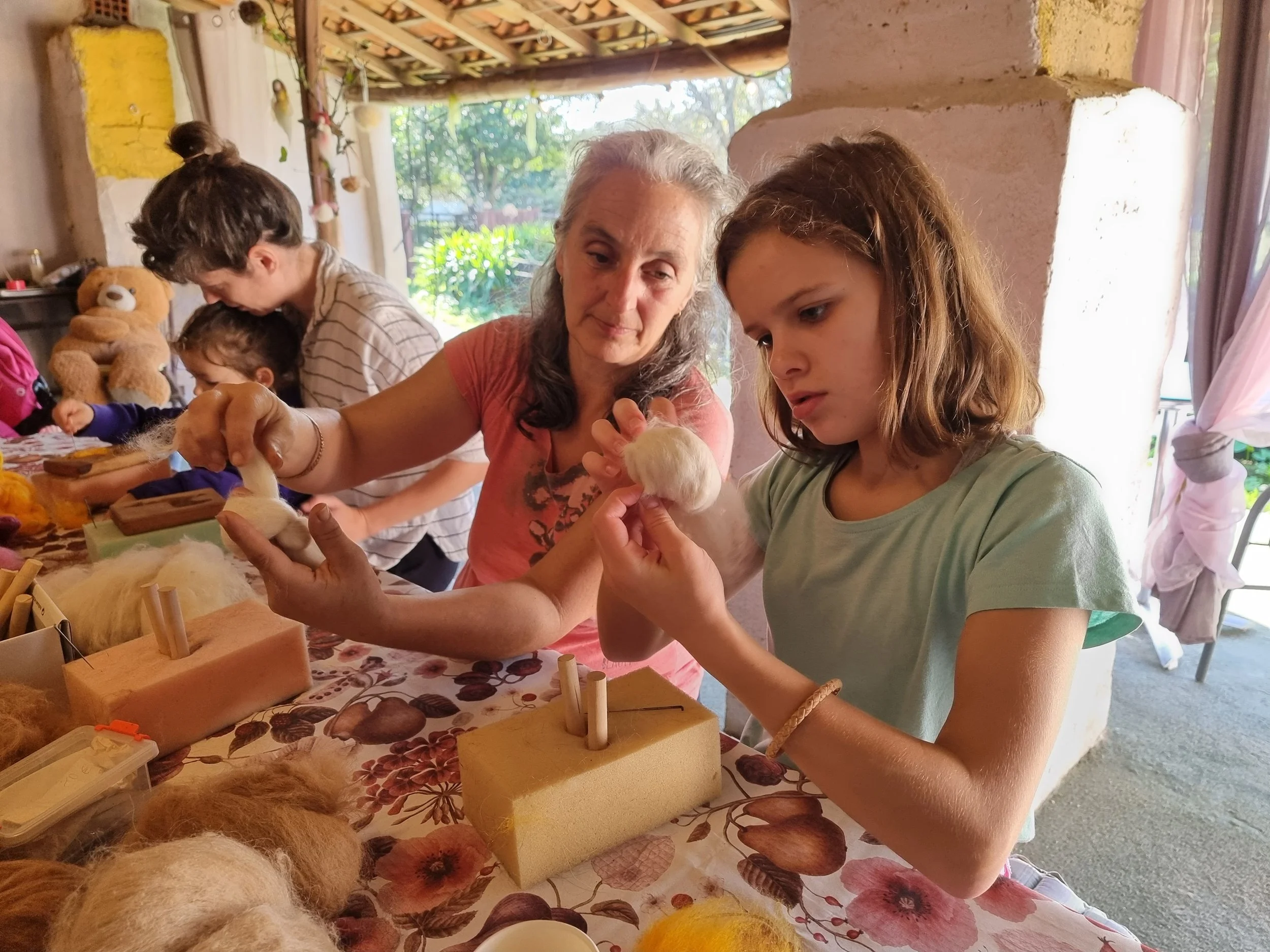 A group of people, including women and children, gathered around a table with craft supplies, working on a felting project at Monte Frio Alpacas. Set in a rustic semi-outdoor space with sunlight and greenery outside.