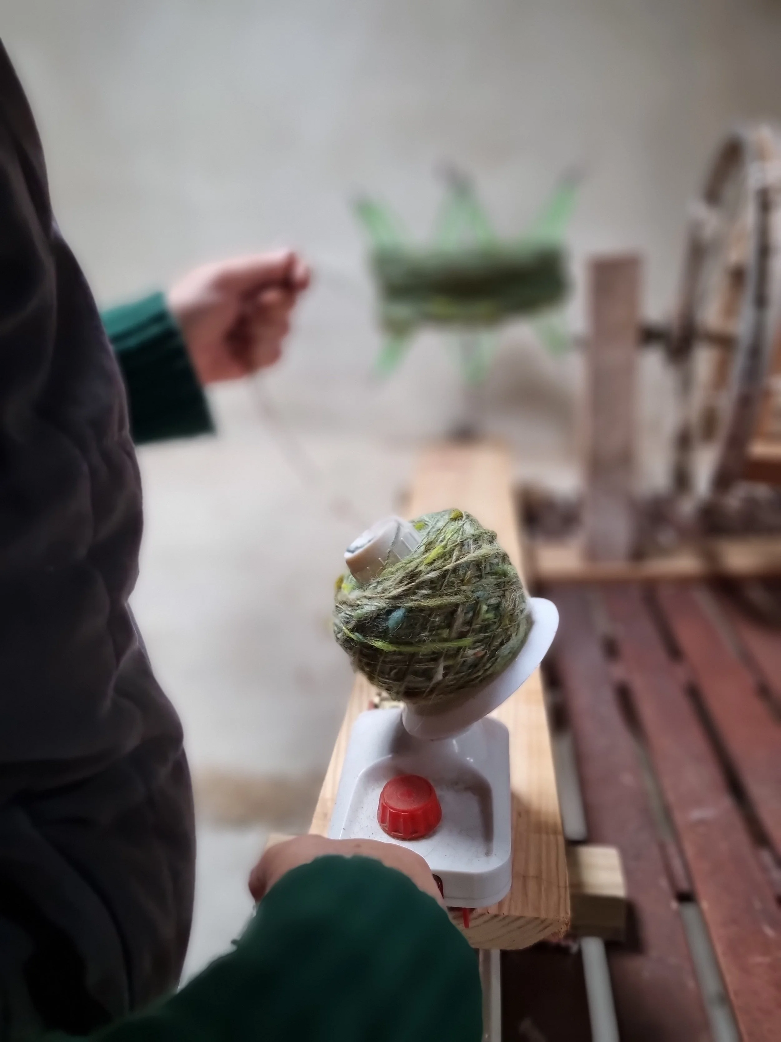 Close-up of a craft project with a small globe wrapped in yarn, held by a person wearing a green and black jacket, standing at a white table.