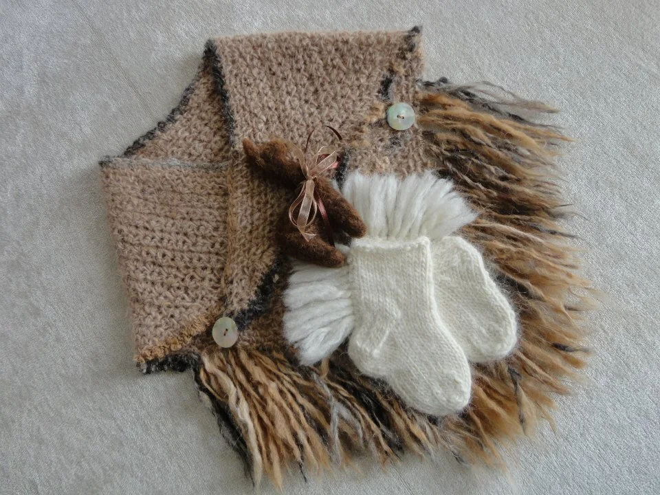 A collection of holiday-themed items arranged on a fluffy, multicolored fur surface. Includes a hand-knit beige scarf with buttons, a small brown plush bear with a ribbon and a pair of cream-colored knit mittens. Made at Monte Frio Alpacas.