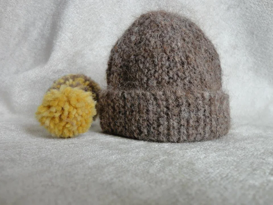 A brown wool knit beanie with a folded brim and a yellow pom-pom, placed on a white textured surface. Made at Monte Frio Alpacas. 