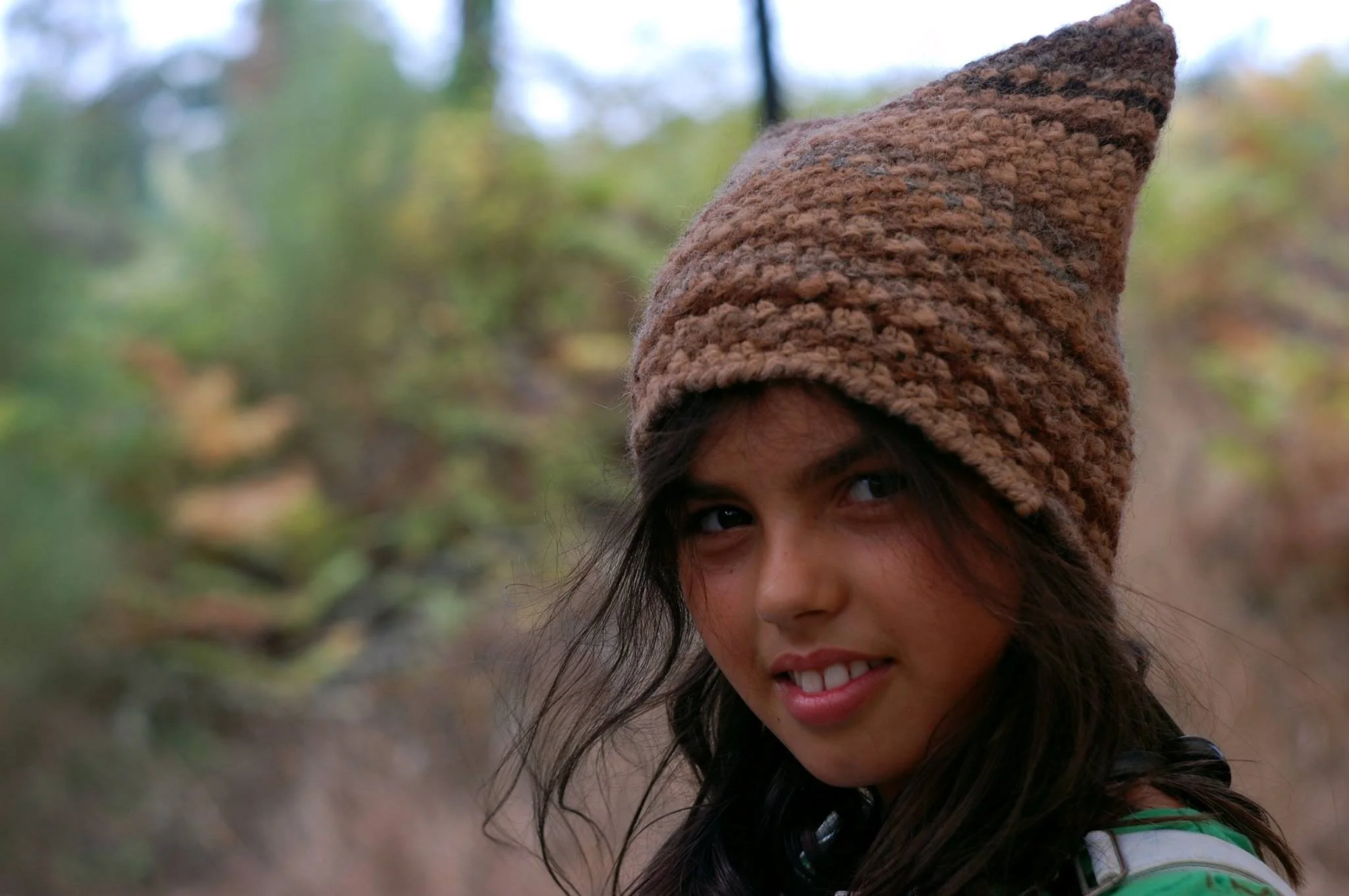 A young woman with dark hair and brown eyes wearing a brown hand knitted hat, outdoors in a natural setting. Made at Monte Frio Alpacas. 