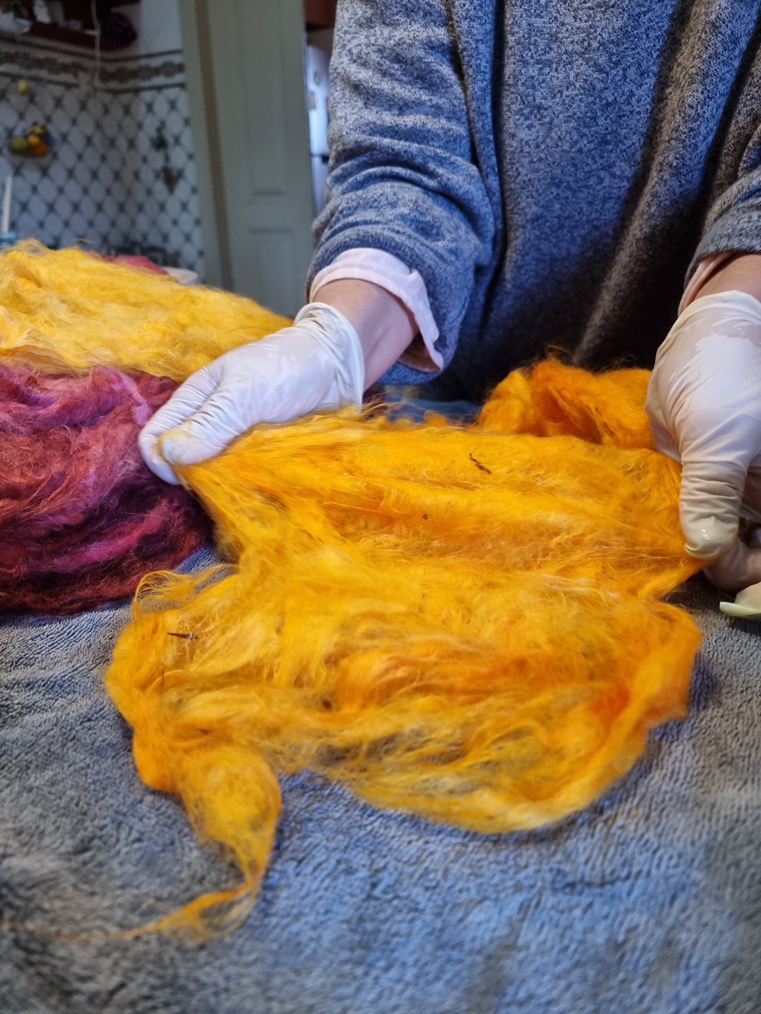 Close-up of a person wearing gloves working with a large, colorful piece of wool, involved in a felting or textile craft project, with a textured gray surface underneath.