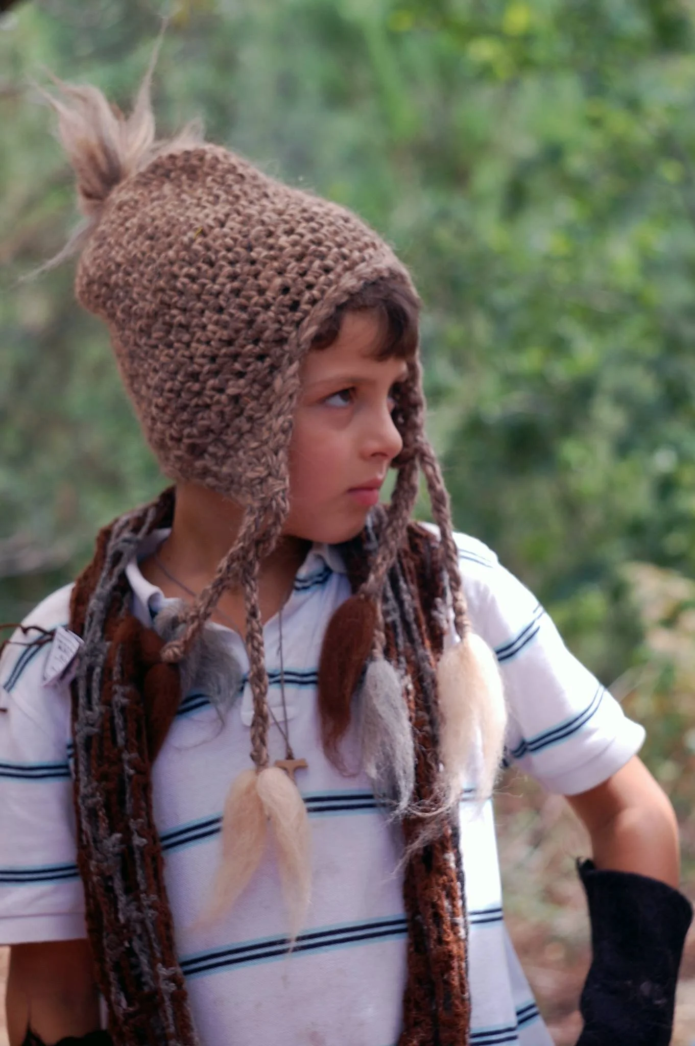 A young boy wearing a brown handmade knit hat with earflaps and pom-poms, and a white shirt with blue stripes, outdoors in a green, leafy setting. Made at Monte Frio Alpacas. 