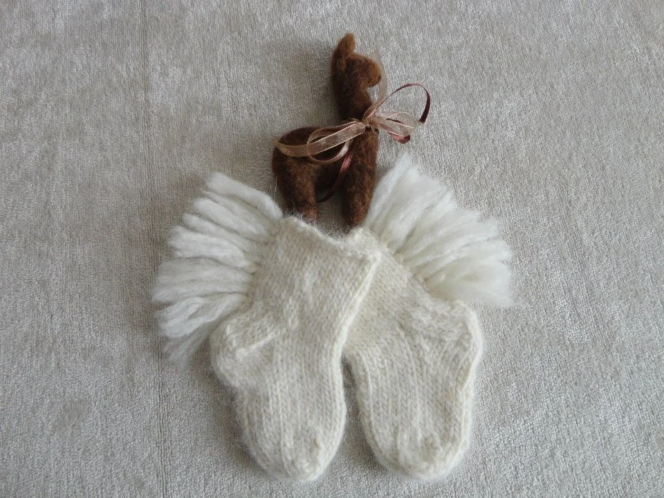 A pair of small white knit baby socks with fringed edges and a tiny brown felted reindeer ornament with a pink ribbon bow, placed on a light-colored fabric surface. Made at Monte Frio Alpacas.