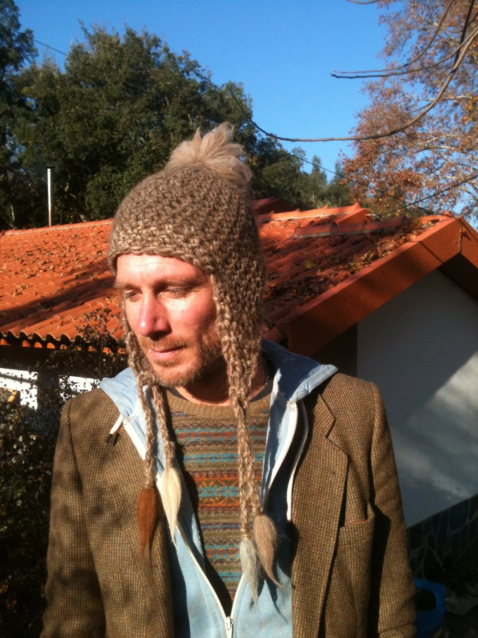 A man with a beard wearing a handmade knitted hat with braids, a brown checkered jacket, and a colorful sweater underneath, standing outdoors in front of a house with a red tile roof and autumn trees. Made at Monte Frio Alpacas. 