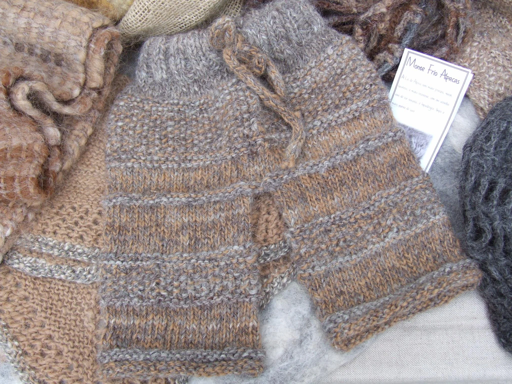 Various handmade knitted and woven woolen scarves and hats in natural and earthy tones displayed on a surface, with a nearby information card. Made at Monte Frio Alpacas. 