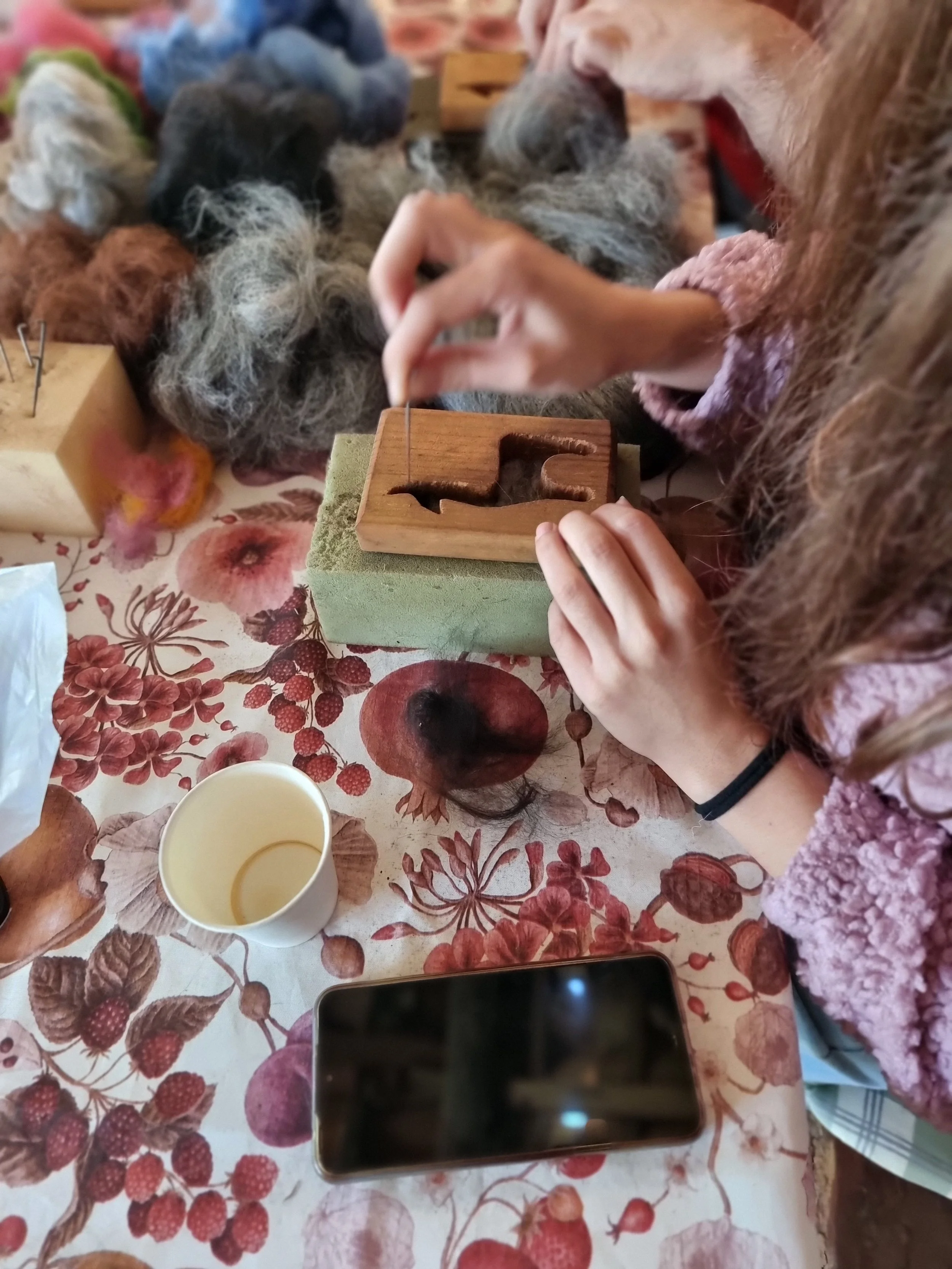 A photo taken during a workshop experience at Monte Frio Alpacas. There are multiple small hand felted animals and fairies in different colors and textures on the table, which has a floral-patterned tablecloth with berries and flowers.