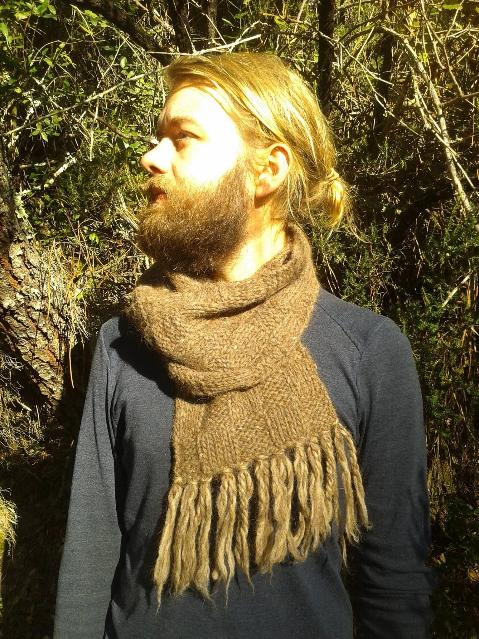 A man with blonde hair and a beard wearing a brown handmade scarf and dark clothing, standing outdoors among dense green bushes and trees. Made at Monte Frio Alpacas. 