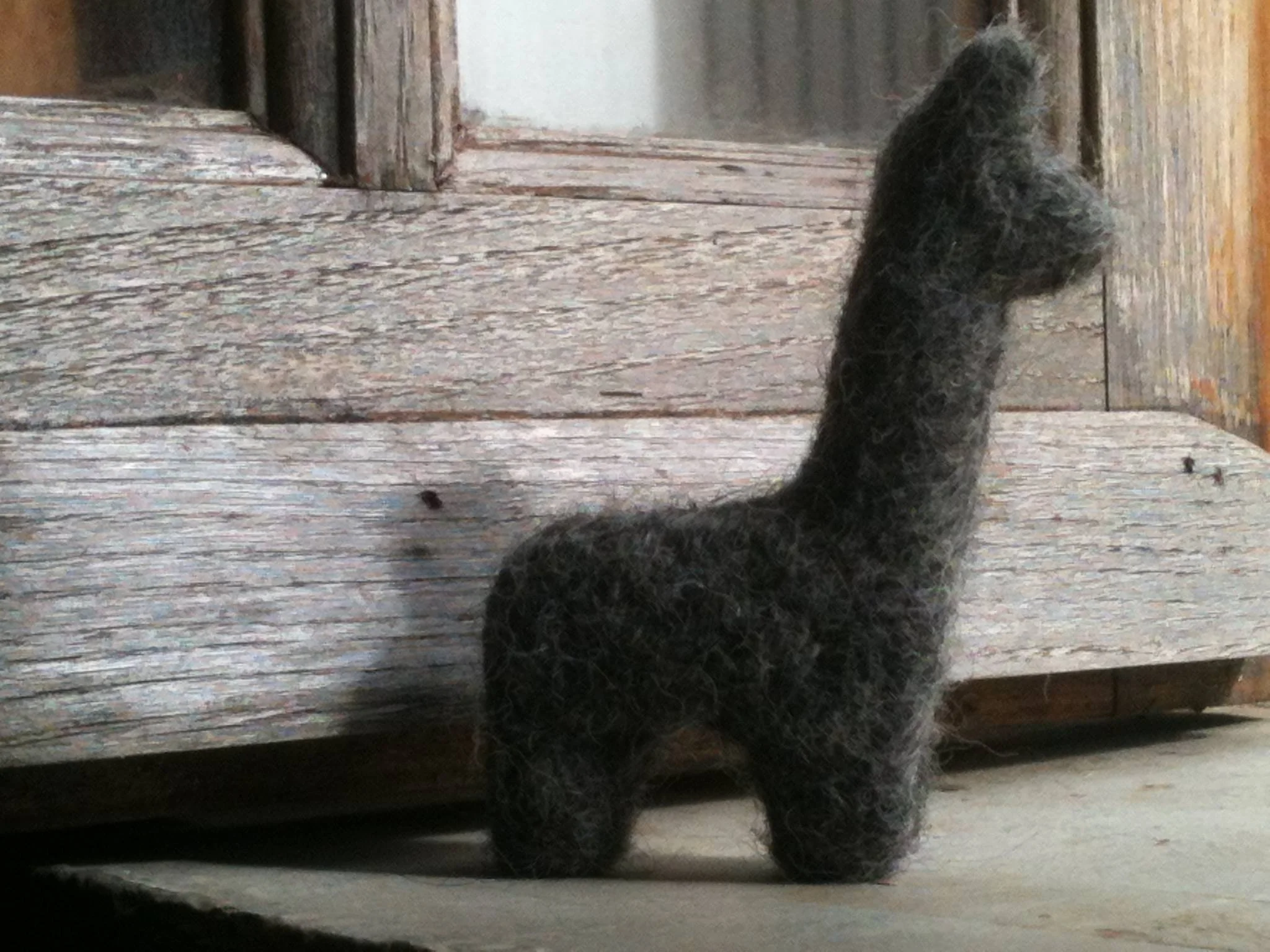The image shows a black hand felted alpaca sitting on a wooden floor. Made at Monte Frio Alpacas.