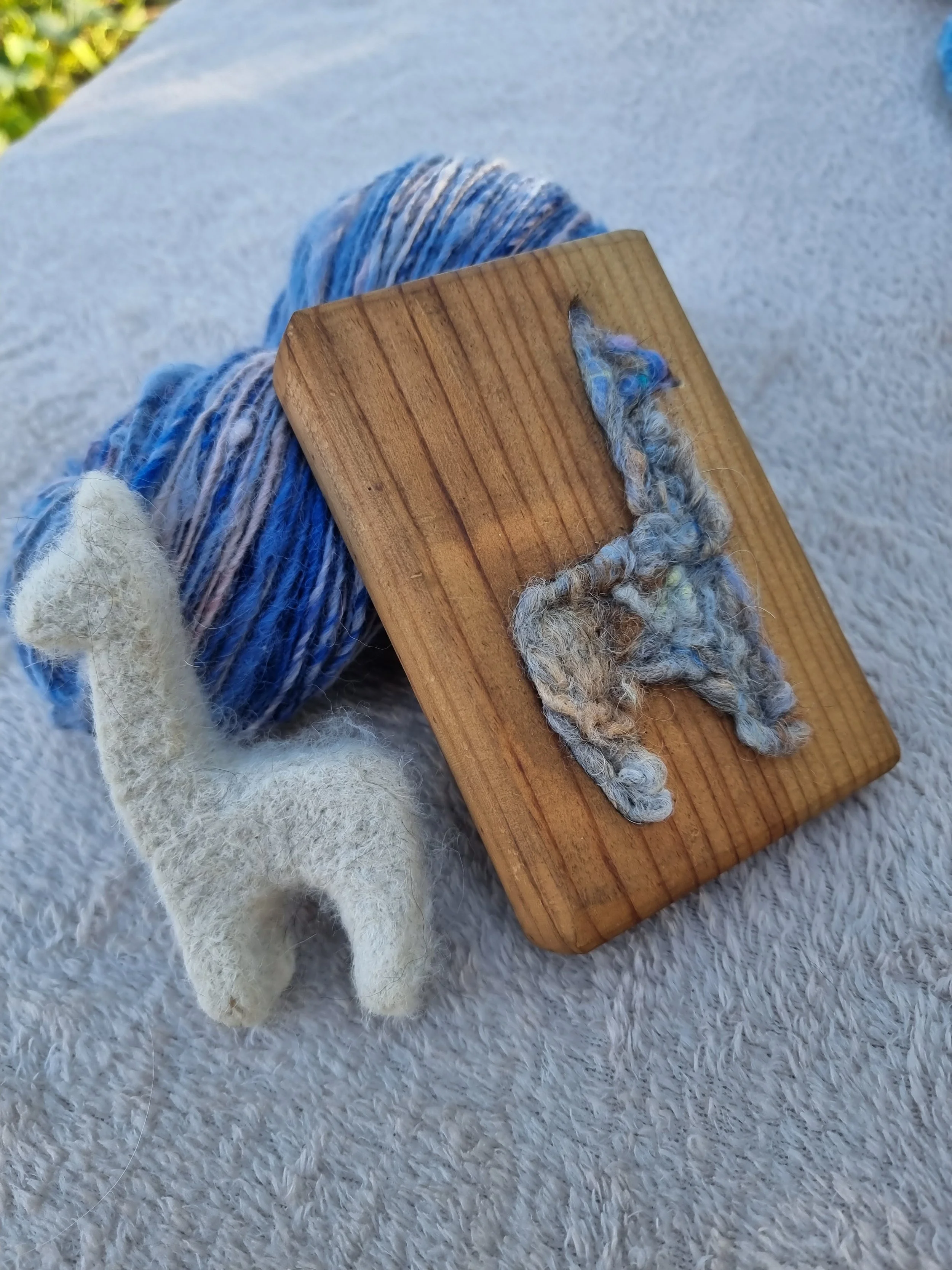 Close-up of a wooden weaving shuttle with blue and white yarn, resting on a fluffy white surface. A hand felted alpaca rests on the wool. Made at Monte Frio Alpacas.