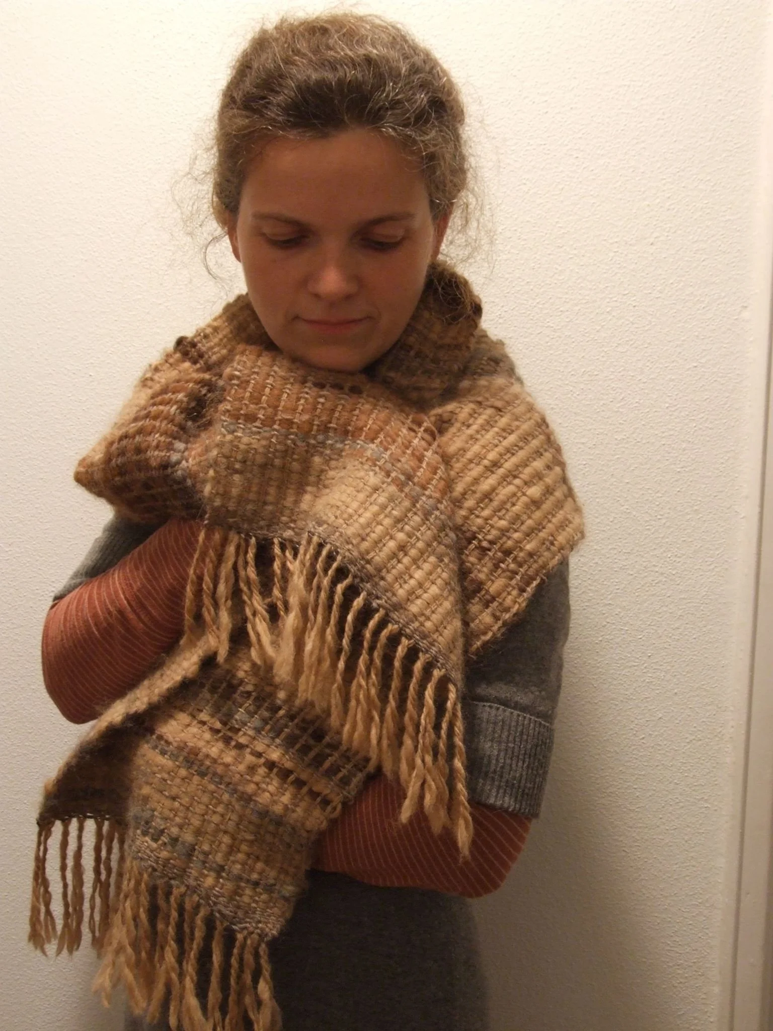 A woman with curly hair wearing a checked scarf, red shirt, and gray sweater standing against a plain wall. Made at Monte Frio Alpacas. 