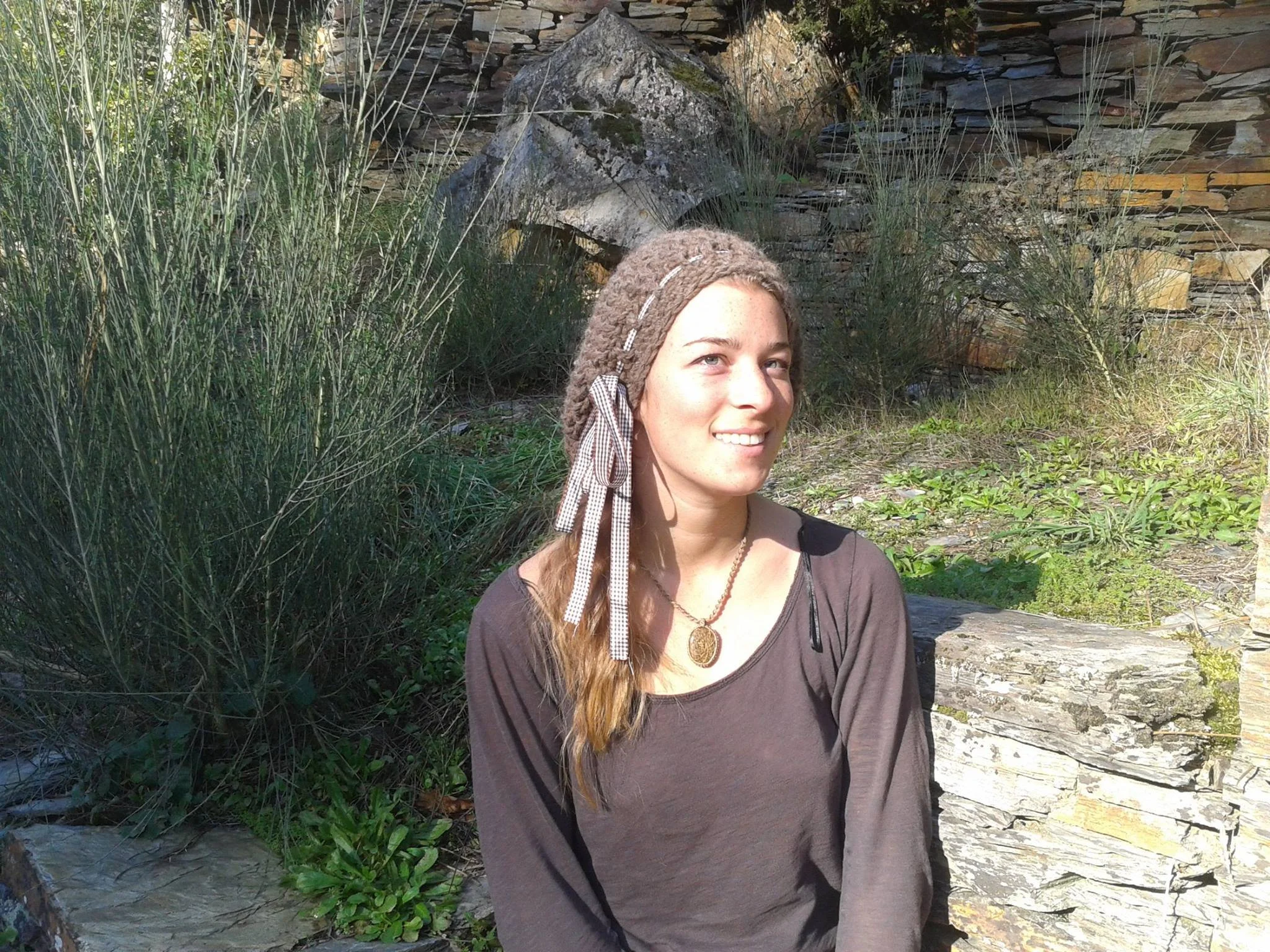 A smiling woman with long, light brown hair, wearing a brown handmade hat and a necklace, sitting outdoors near a stone wall with plants and grass around her. Made at Monte Frio Alpacas. 