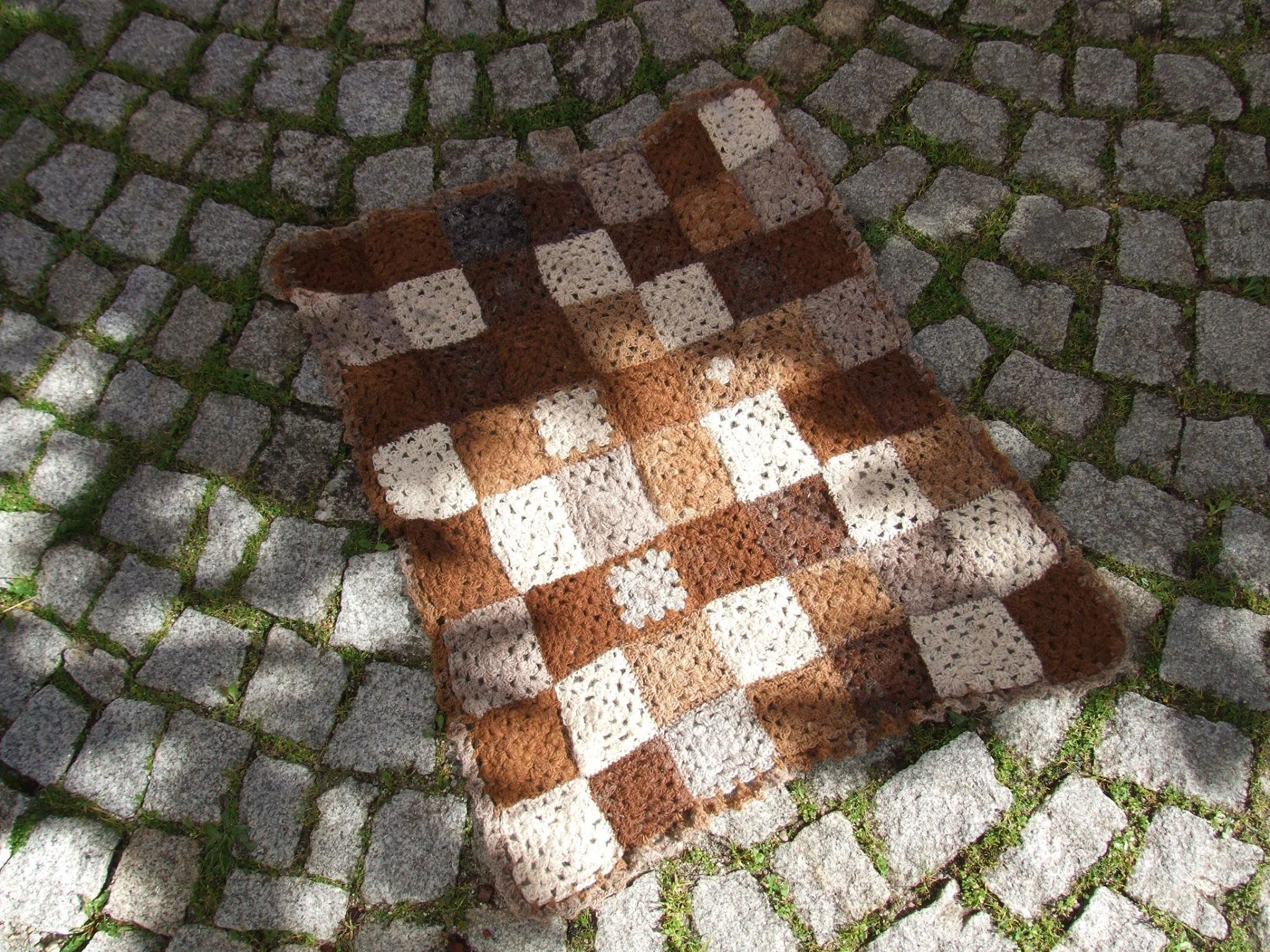 A handmade crocheted blanket with a checkered pattern in shades of brown, beige, and cream, lying on a cobblestone pathway surrounded by grass. Made at Monte Frio Alpacas.