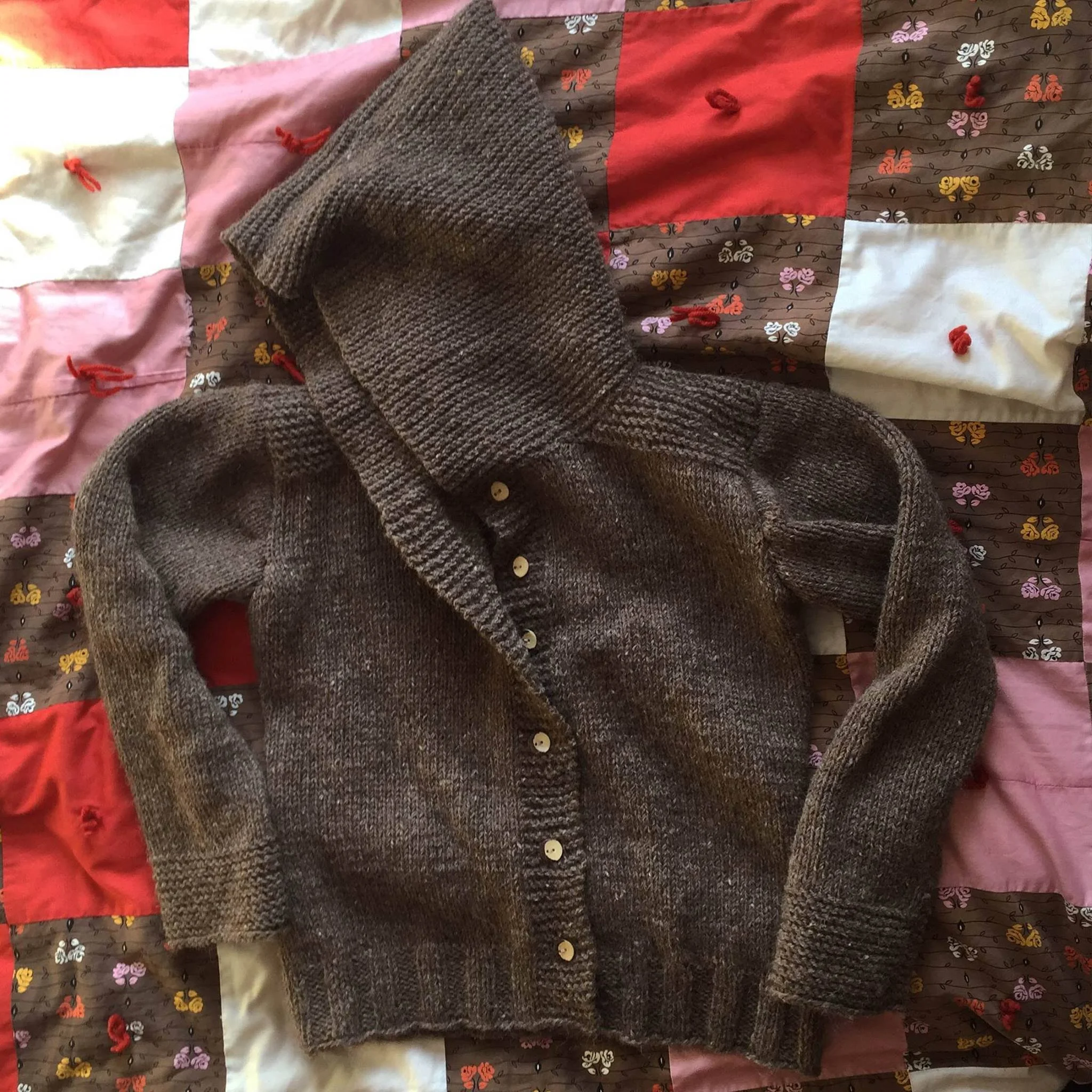 A folded gray knitted cardigan with buttons resting on a colorful quilted bedspread with pink, white, red, and brown patches with floral patterns. Made at Monte Frio Alpacas. 