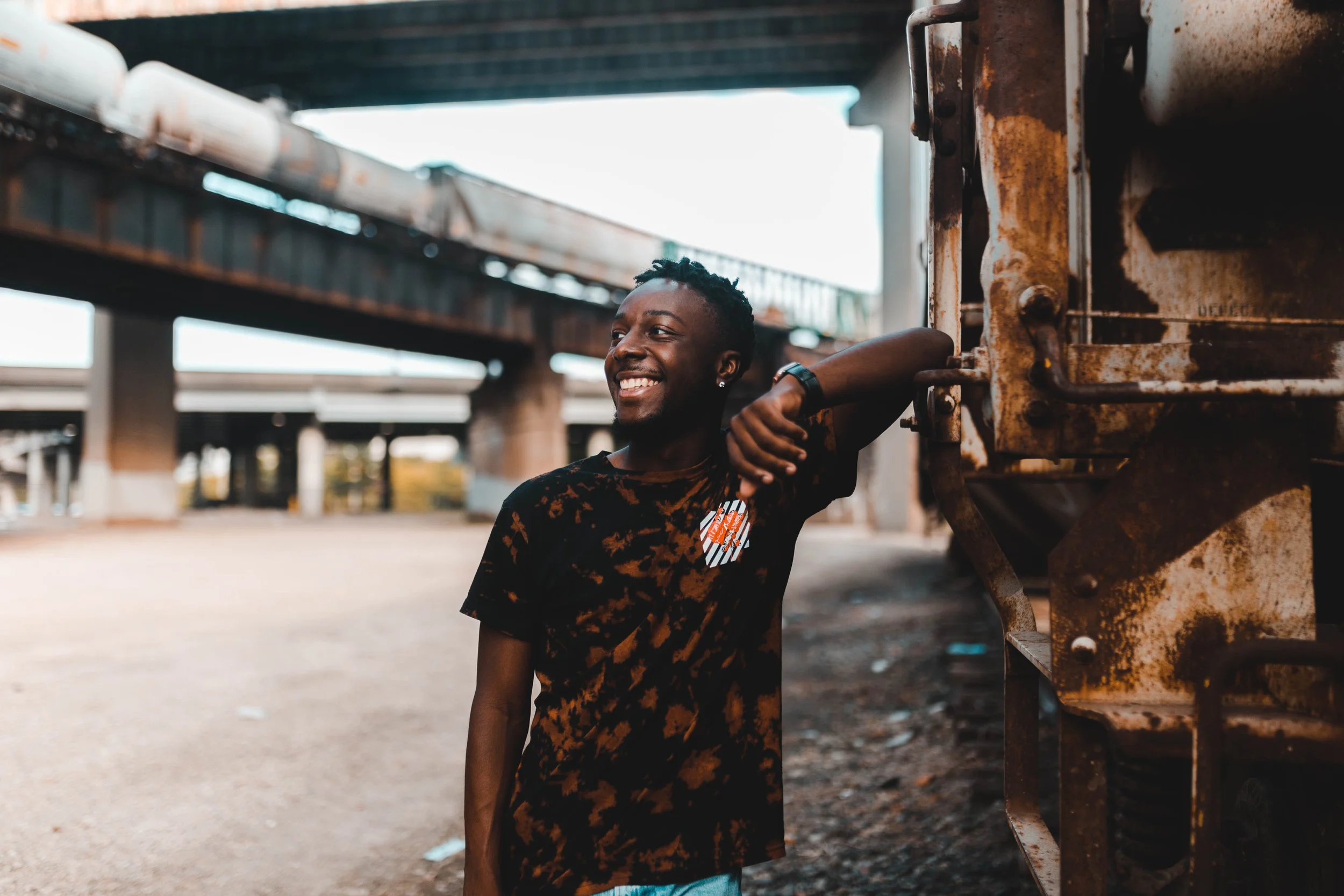 A smiling young man leaning on a rusty train car under an overpass in an industrial area.