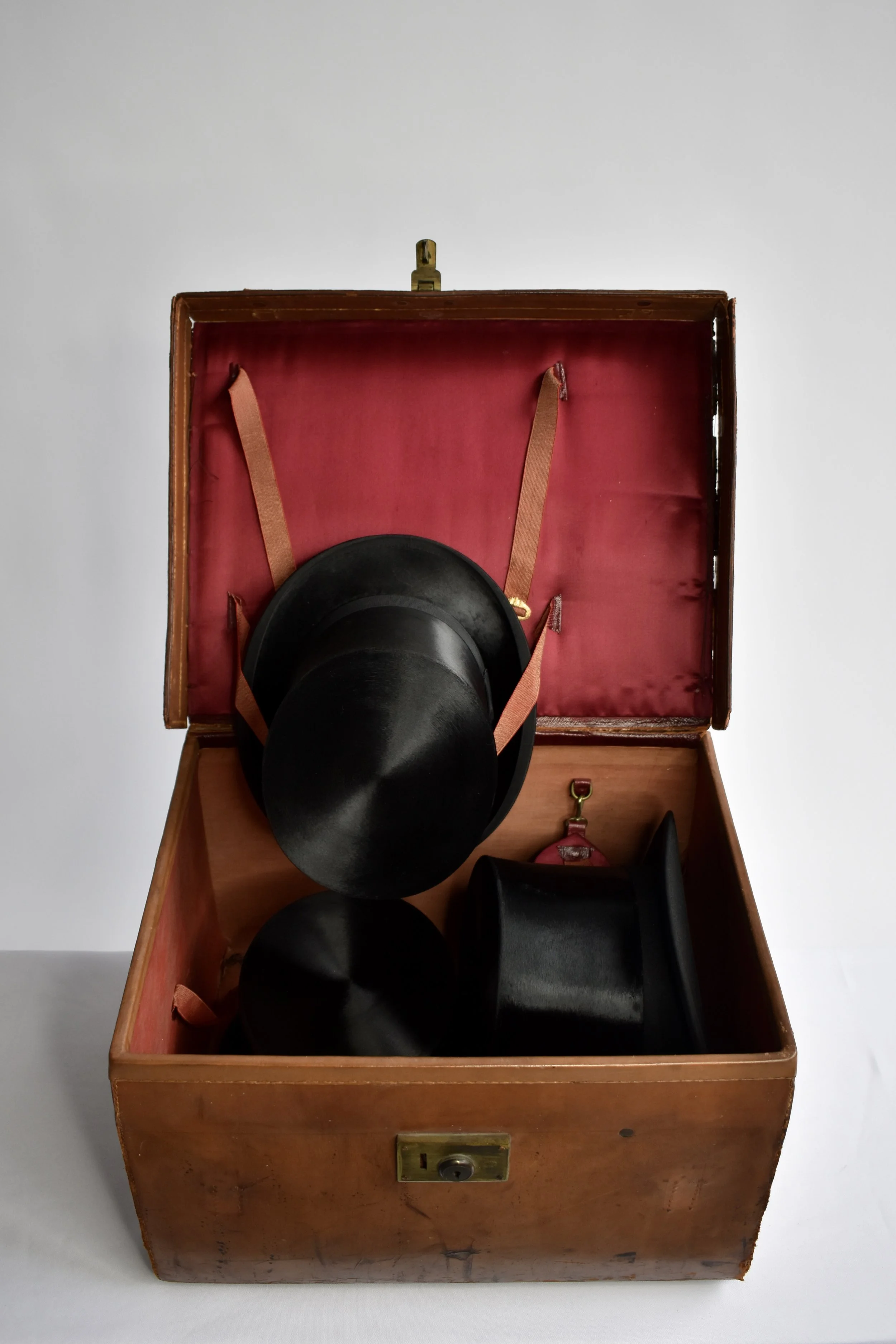 An open wooden box with a red interior containing three black top hats, with a brass latch on the front.