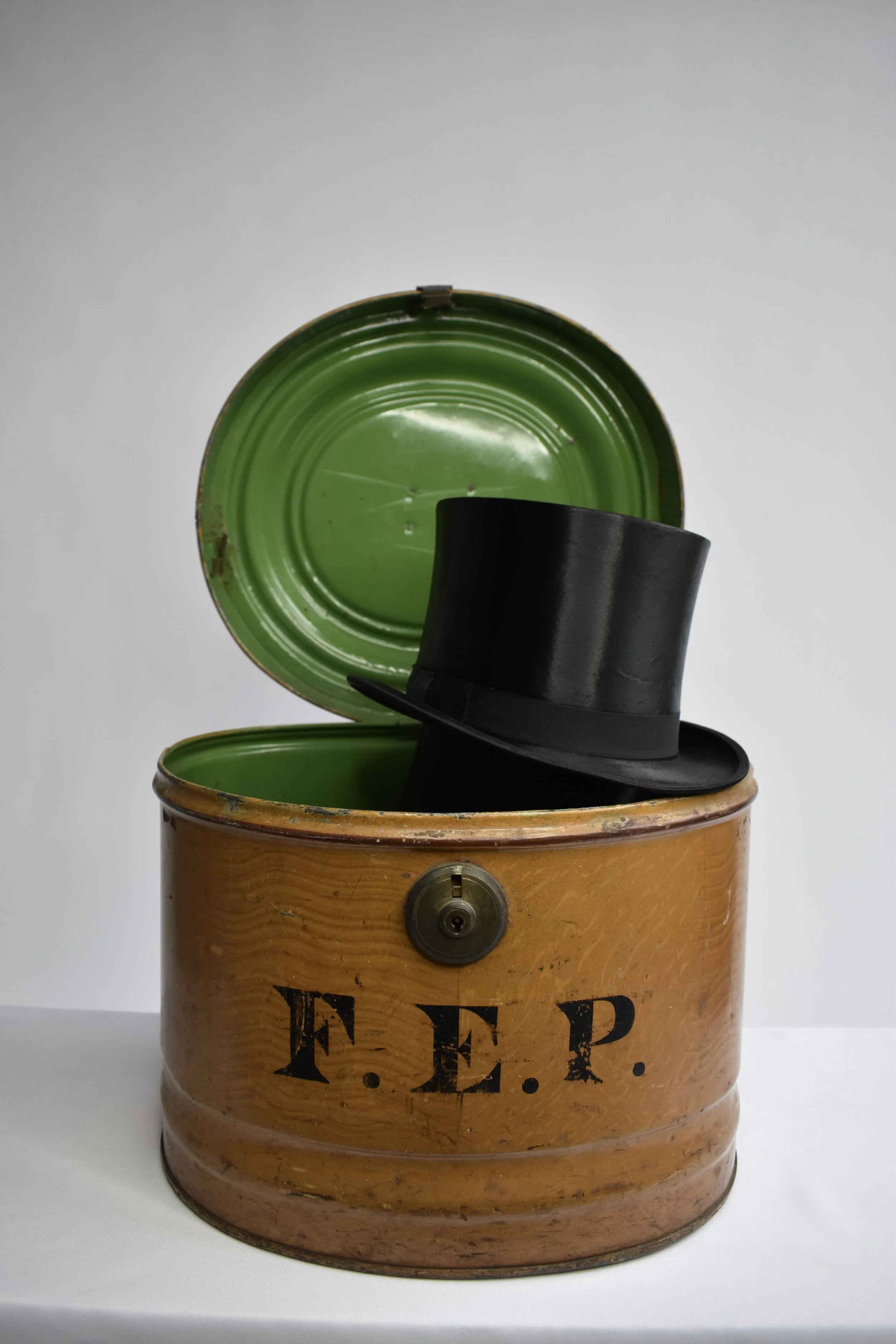 An open vintage tin with a painted green interior vintage lock, labeled "F.E.P." in black stencilled letters, with a black silk top hat resting on top.