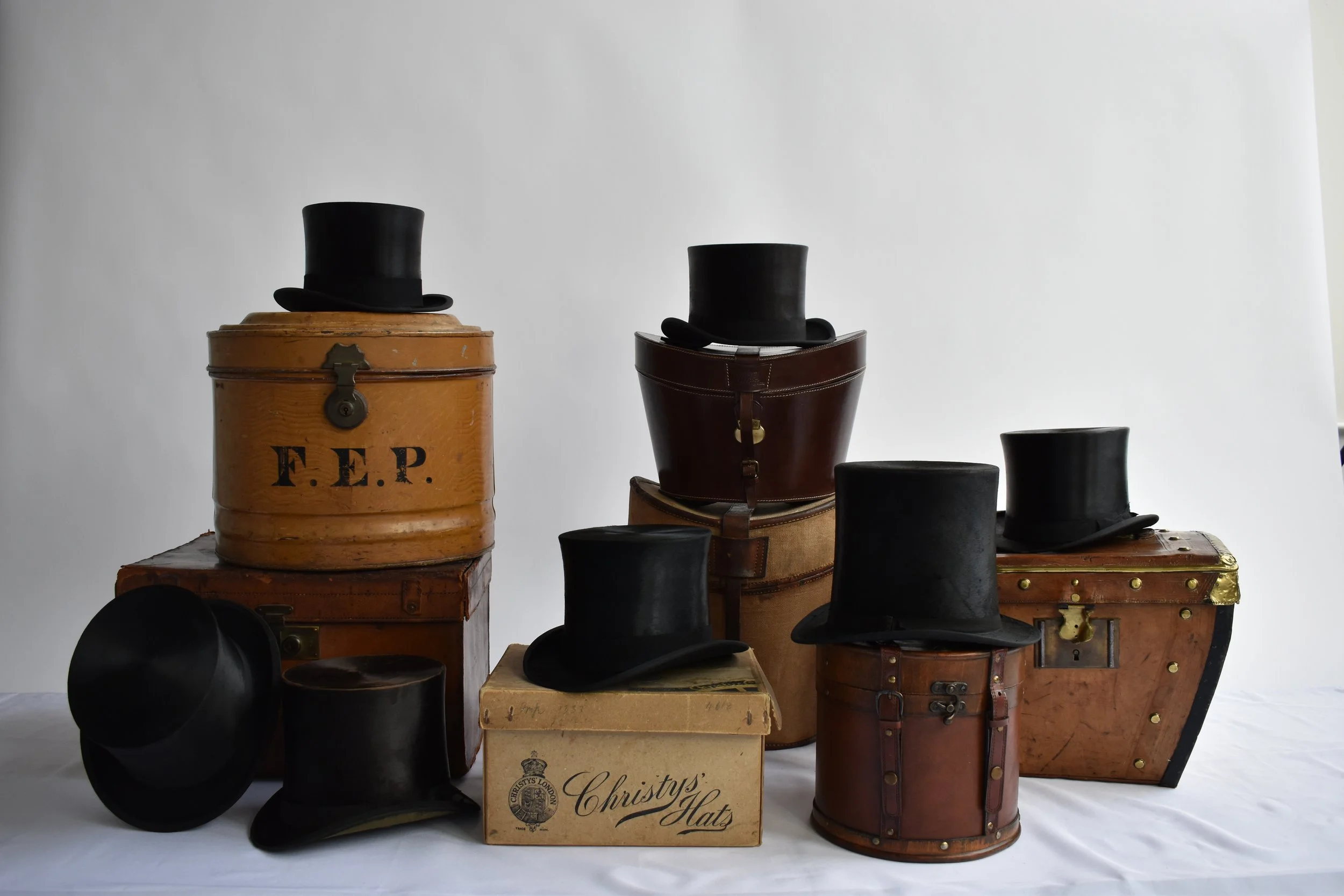A collection of vintage top hats, some on wooden boxes and trunks, with a sign reading 'Christys Hats' in the foreground, set against a plain white background.