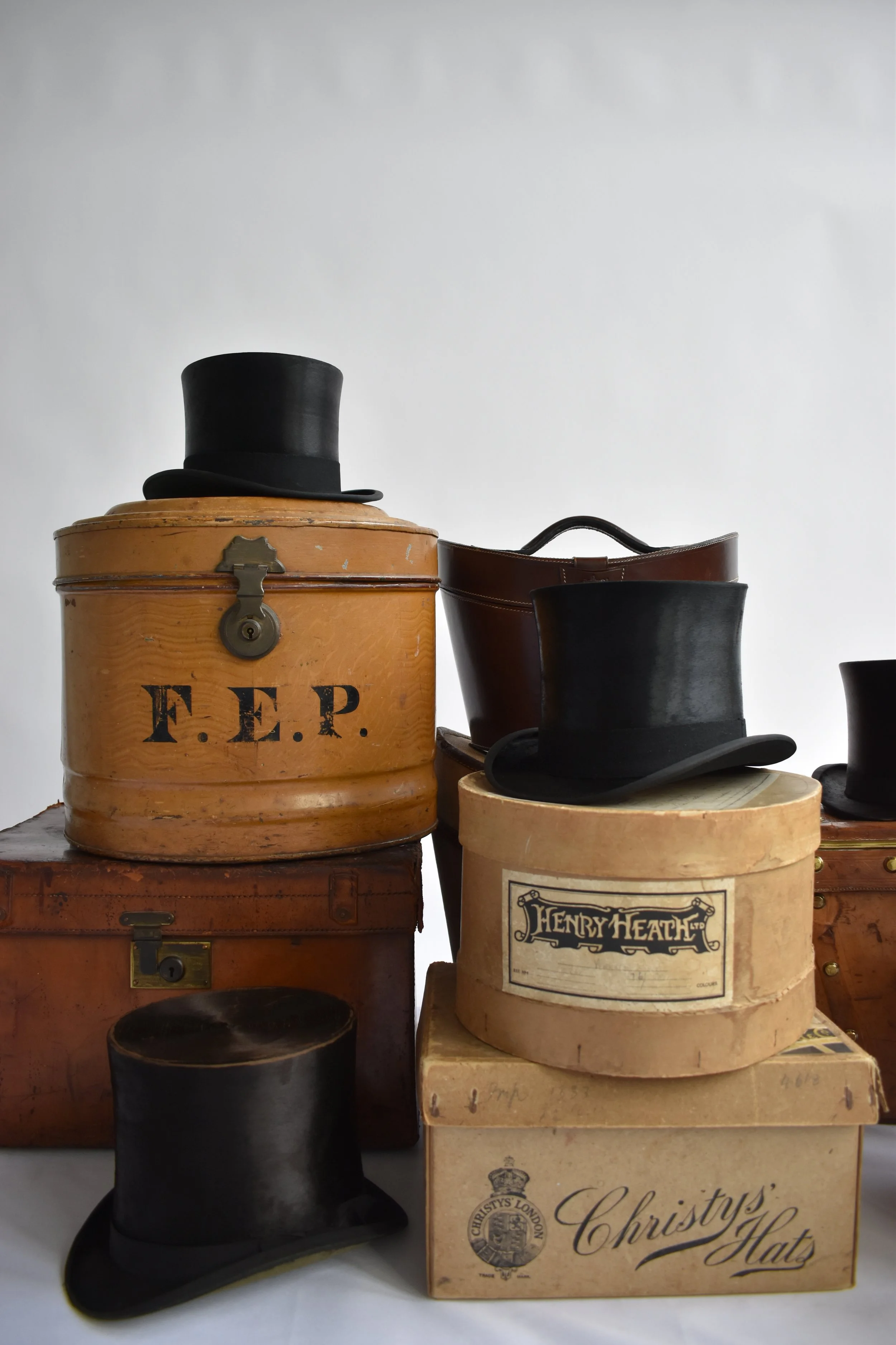 Various vintage hats, including top hats and bowler hats, are displayed on top of their boxes and cases, some labeled with brands like Christy's.