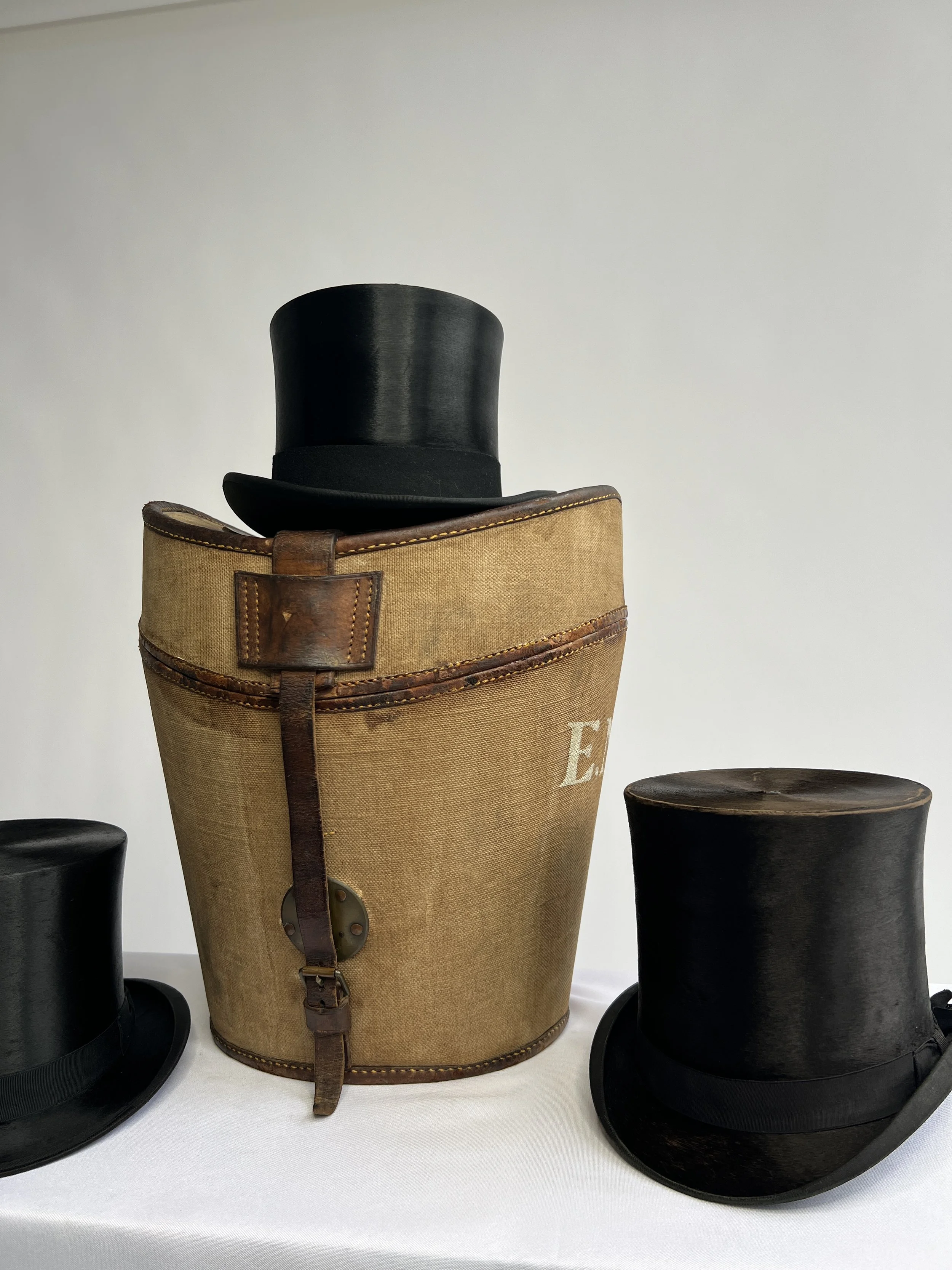 Three black top hats and a vintage tan costume hat with leather straps on a white surface.
