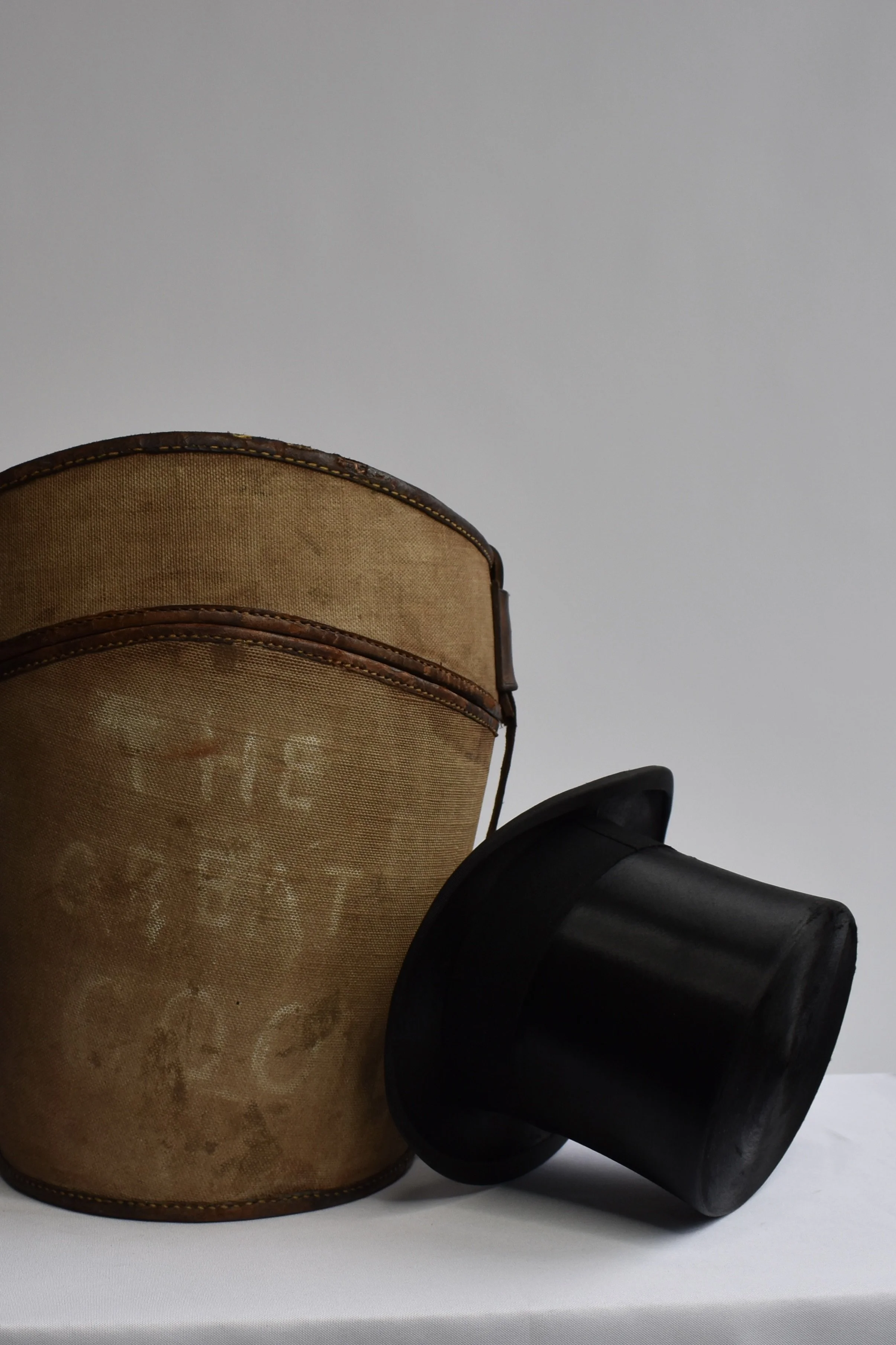 Close-up of a vintage brown leather and fabric drum with a black drumstick resting on a white surface against a plain gray wall.