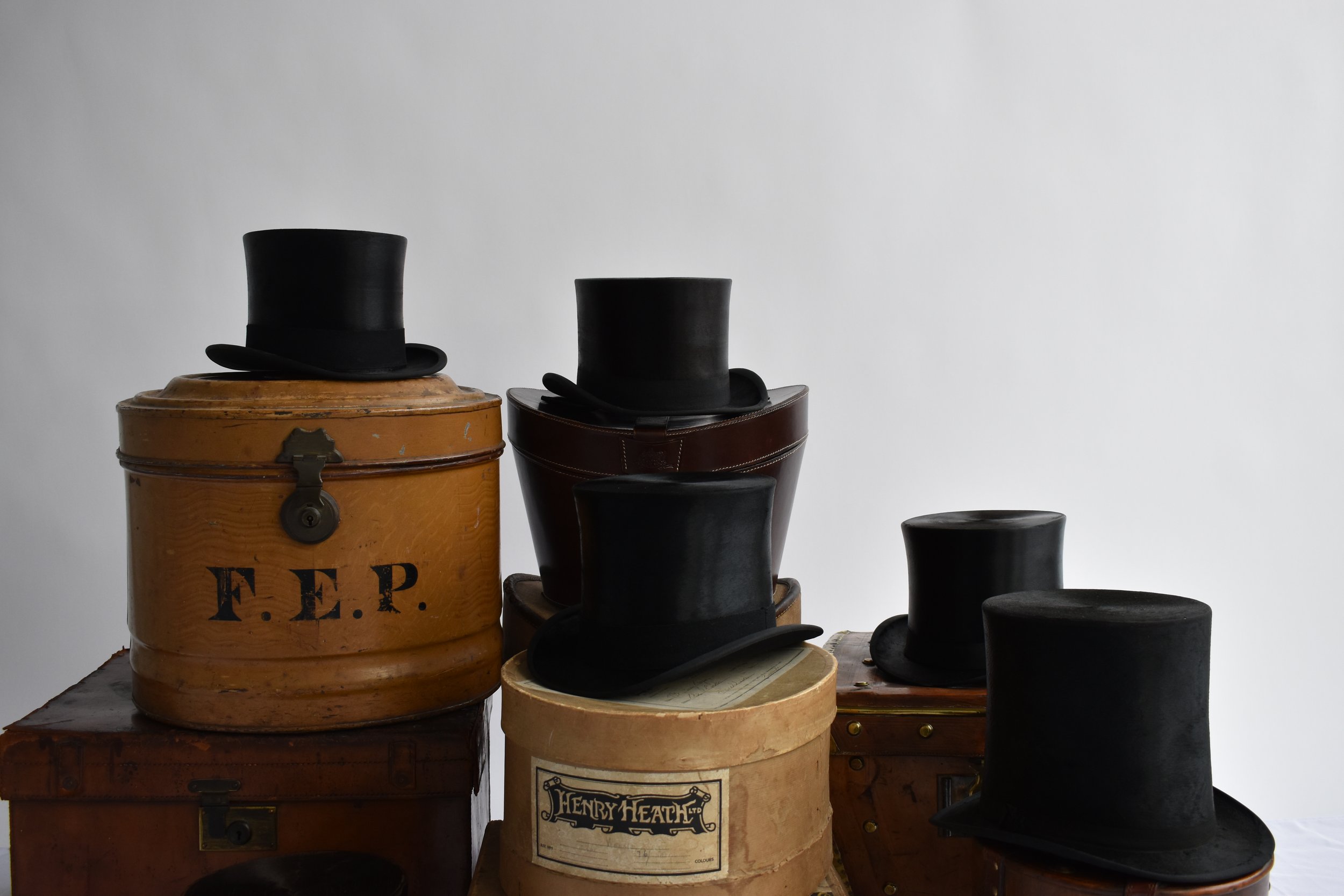 Several vintage black top hats and a bowler hat on top of wooden and leather boxes, with a plain light background.