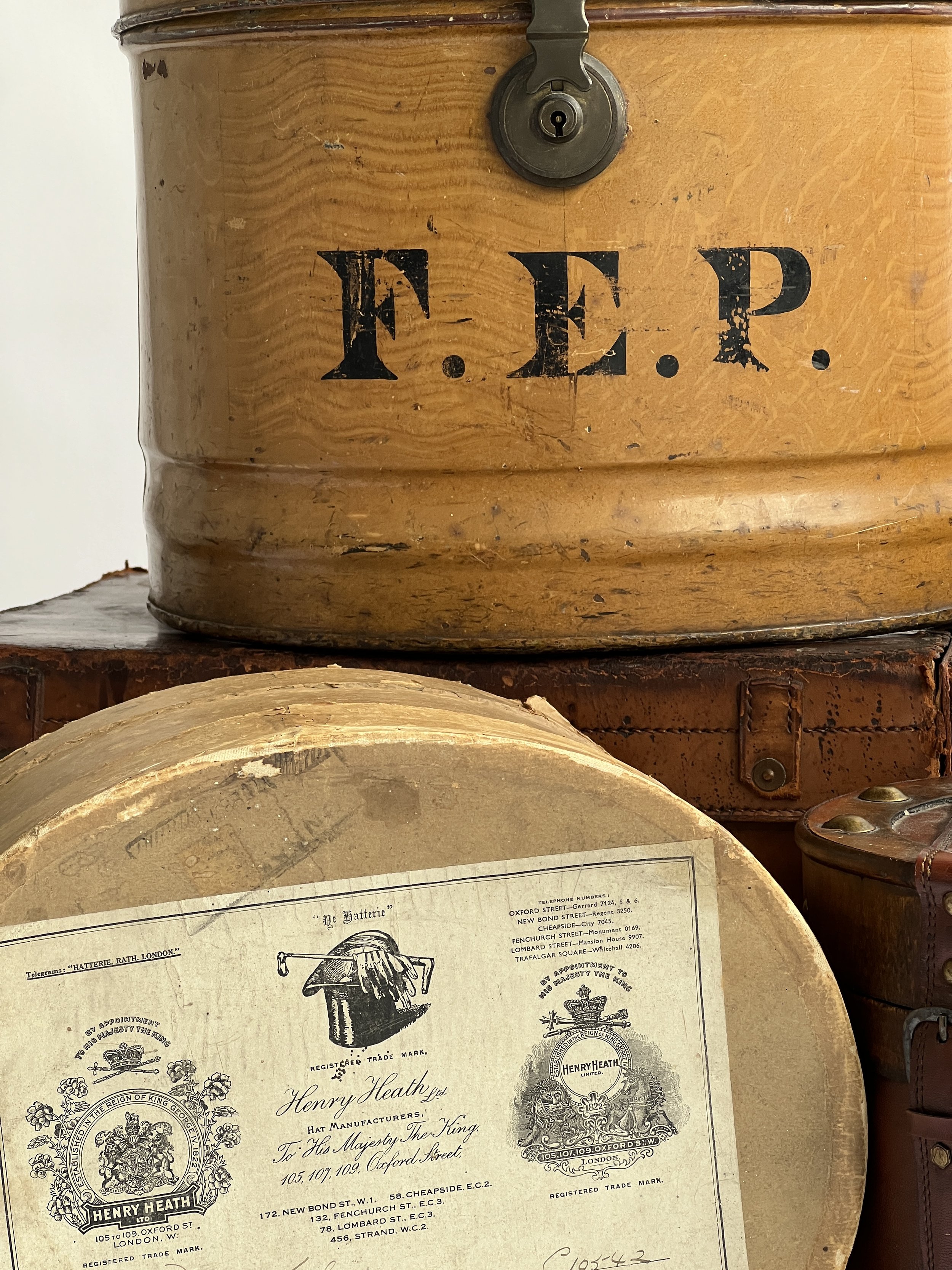 Close-up of a vintage wooden box with a metal lock and the initials 'F.E.P.' painted in black. Below, part of an old document or label from Henry Heath Ltd., a hat manufacturer, with vintage typography and detailed crests.