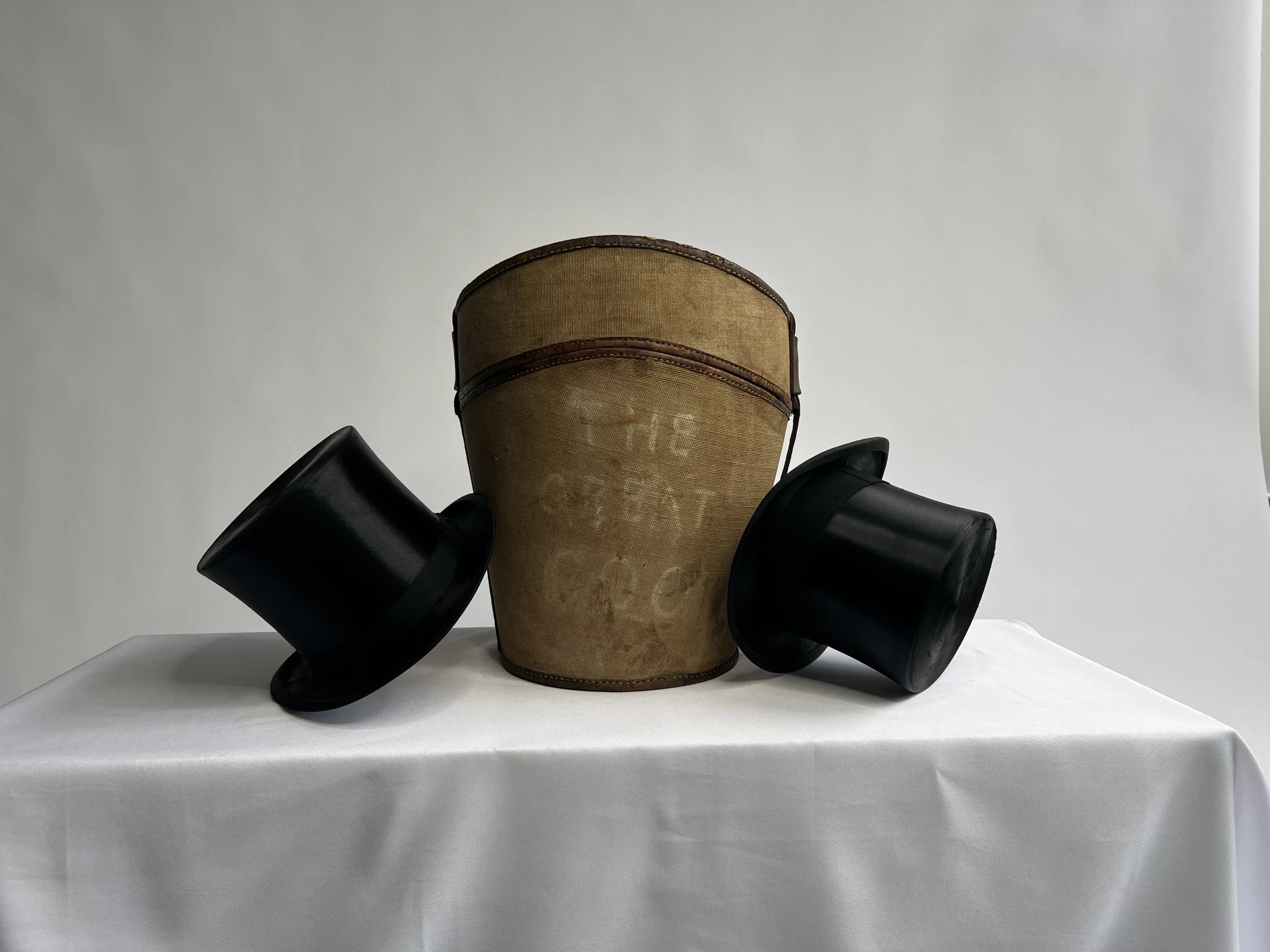 A vintage light brown canvas hat case hat with a wide brim, photographed with black two silk top hats.